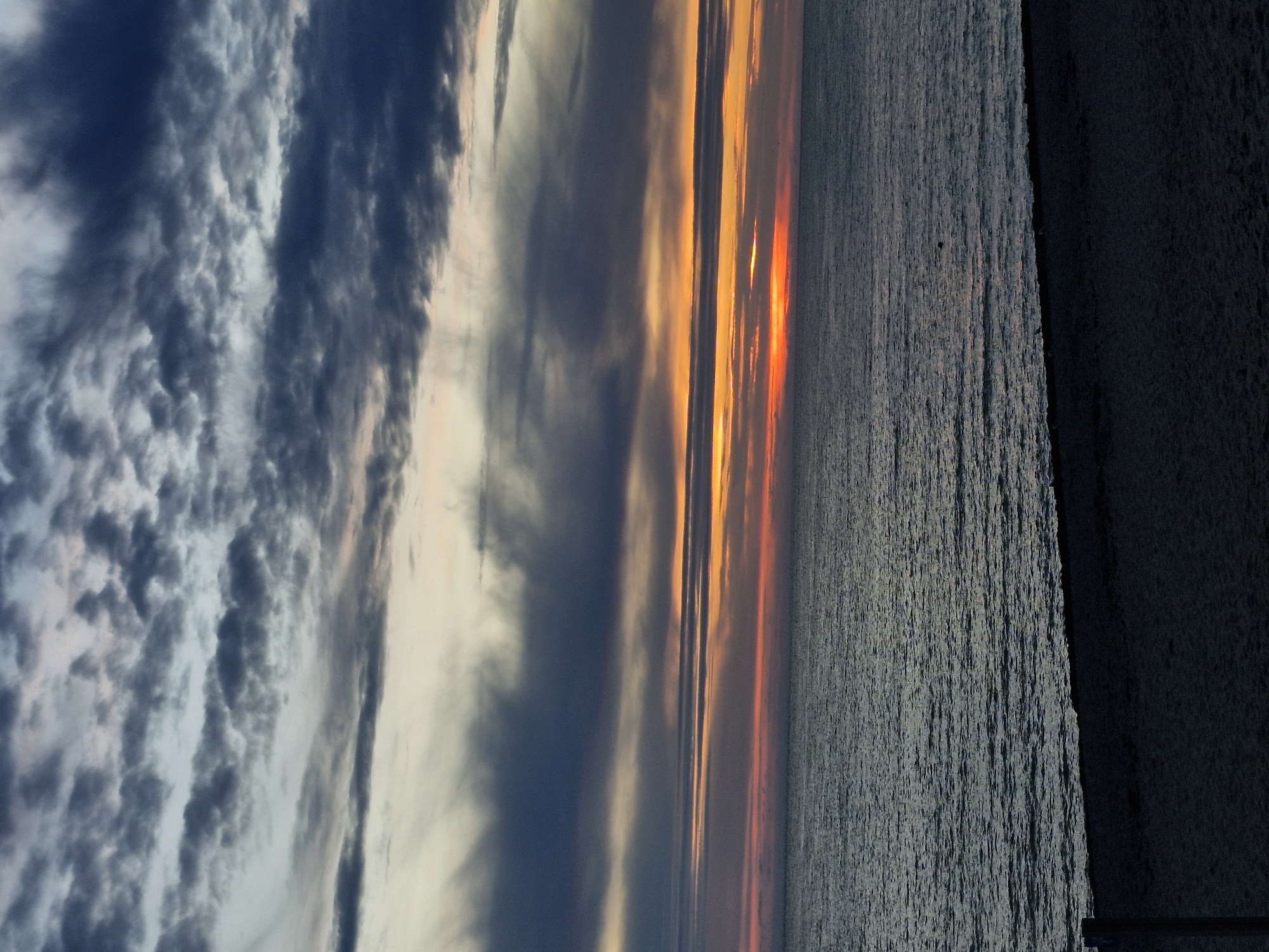 This is a photograph of a sunset over the Baltic Sea, likely taken from the beach in Klaipėda, Lithuania, near the Smiltynė ferry terminal or the Curonian Spit. The sky is filled with dramatic, dark grey and bluish clouds, streaked with thinner bands of orange and pink where the sun is setting. The sea is a deep, muted blue-grey, with gentle ripples reflecting the colors of the sky. In the foreground, a dark, silhouetted beach stretches across the bottom of the frame, appearing sandy and empty except for what might be a faint wooden post on the far left. The overall mood is serene and somber, characteristic of a cloudy sunset.
