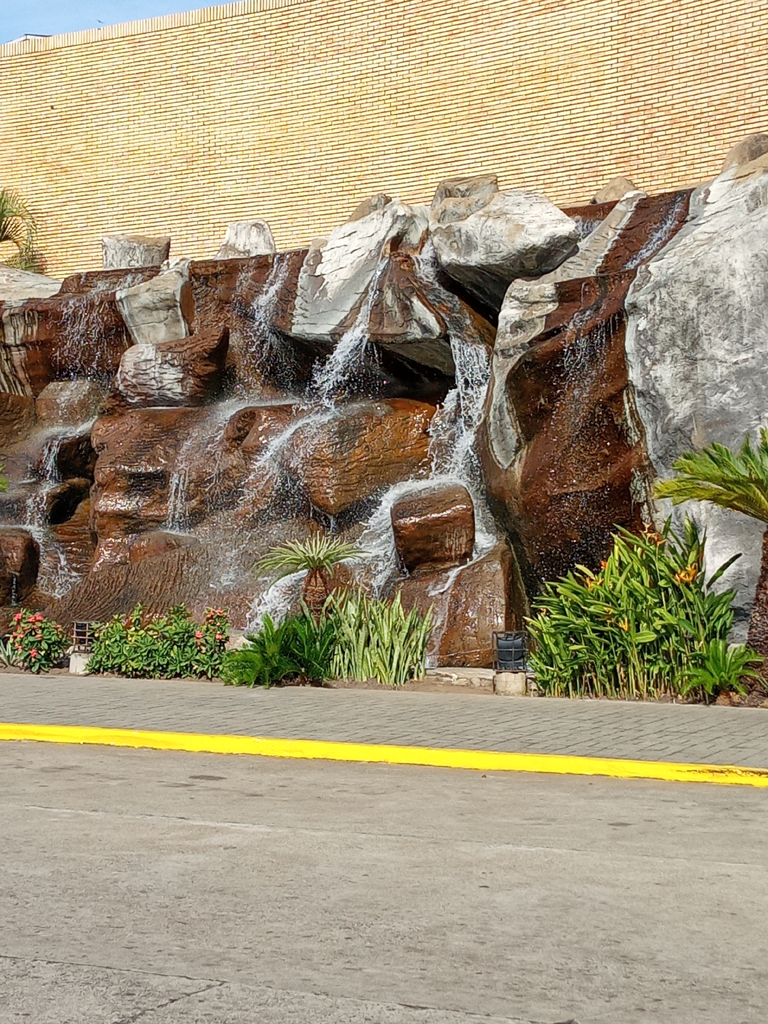 The image depicts an artificial waterfall cascading over rocks of brown and gray hues, positioned in front of a building with a brick facade. The base of the waterfall is surrounded by green plants and flowers. A yellow line marks the edge of a paved area in the foreground. The location is Guayana City, Venezuela. No people are visible.