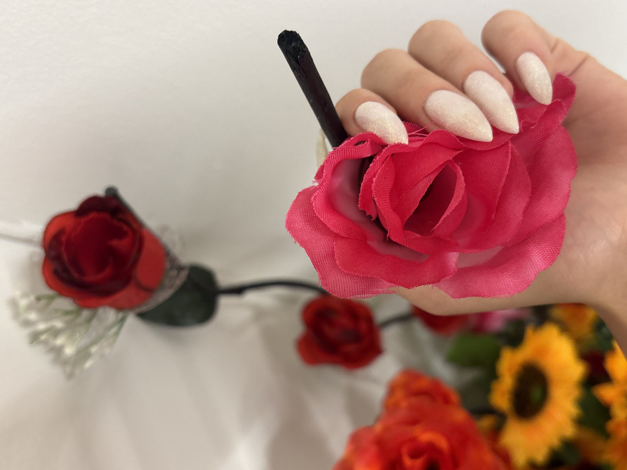 In a shallow depth of field, a hand with pale, glitter-infused stiletto nails holds a vibrant pink artificial rose with a black stem. The rose is positioned in the foreground and is the primary focus. In the blurred background, a red artificial rose with white baby's breath is visible, attached to a dark stem. Further in the background, other out-of-focus artificial flowers, including red poppies and sunflowers, can be seen. The overall setting appears to be an indoor environment with a plain white background, possibly a studio or a decorated space. There are no discernible elements that place this scene specifically in Pleven, Bulgaria.