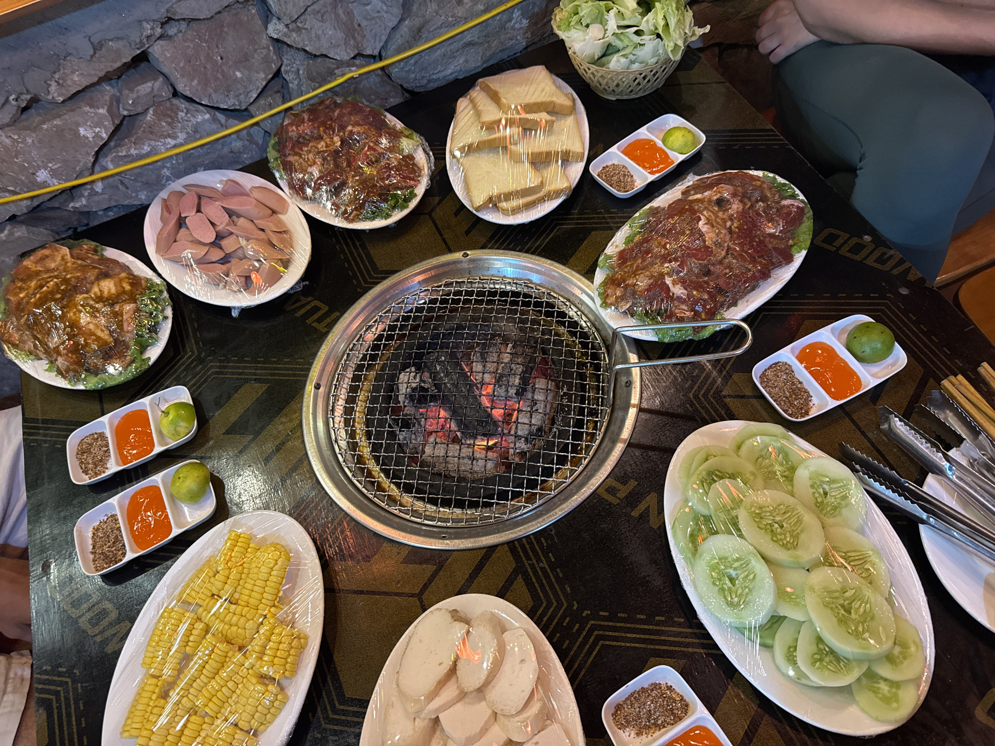 A table at a restaurant in Mèo Vạc Commune, Vietnam, laden with food prepared for grilling. The centerpiece is a charcoal grill, surrounded by plates of raw meat (marinated beef and pork), sliced cucumbers, corn on the cob, sliced fish cakes, sausages, and bread. Small dishes contain condiments like chili sauce, lime wedges, and spice mixes. Utensils, including tongs and chopsticks, are on the table. A basket of lettuce is also visible. A person's legs are seen near the upper right corner of the table. The backdrop features a stone wall. All food items are wrapped in plastic.