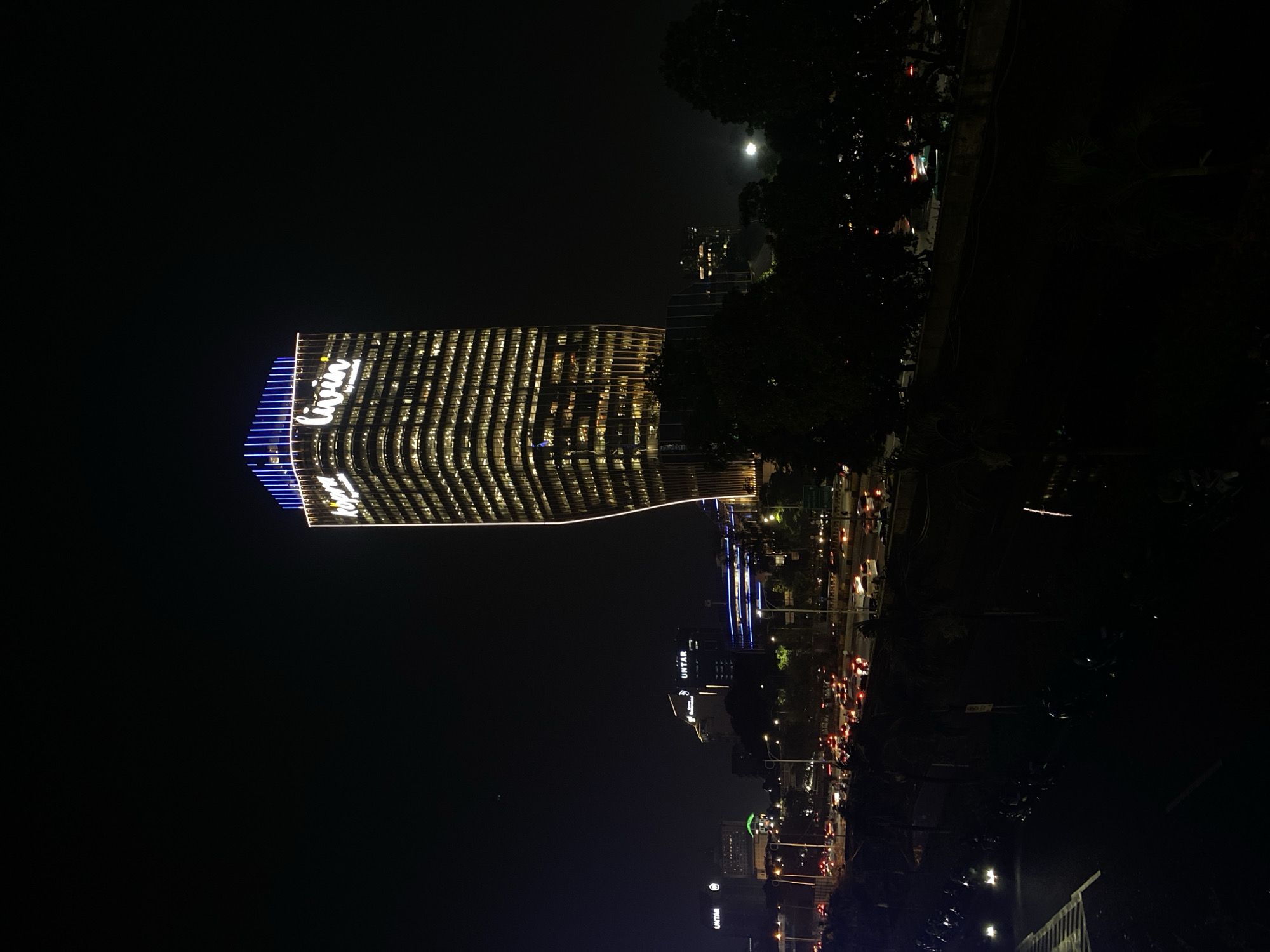 This is a nighttime, slightly rotated, vertical image of a modern, illuminated skyscraper in West Jakarta, Indonesia. The building, with its distinctive wavy facade, is lit from within, showcasing rows of brightly lit windows that create a dynamic pattern. The word "livin" is prominently displayed in white neon lettering across the upper part of the building, accented by blue lights along the edge.
The scene depicts an urban environment at night, with a full moon visible in the dark sky above the trees on the right side of the frame. In the background, other buildings and city lights are visible, suggesting a bustling cityscape. A busy street with car headlights and taillights is also apparent, indicating ongoing traffic. The overall mood is one of urban vibrancy and activity at night. The image is characterized by strong contrasts between the brightly lit building and the dark night sky and surrounding foliage.