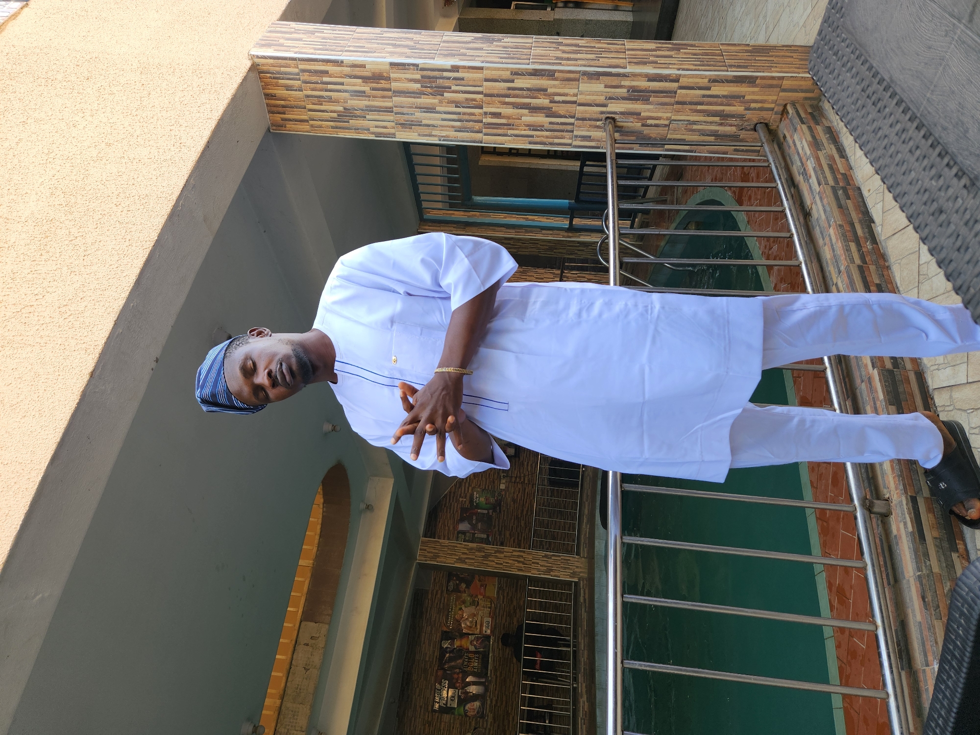 A full-length shot of a man, dressed in a white outfit and a blue head covering, stands with his hands clasped in front of him. He is wearing a gold bracelet on his right wrist and black sandals. The location appears to be an outdoor area with tiled walls and a metal railing. The presence of architectural elements and what looks like an empty pool can be seen behind him. Details such as the texture of the building and the man's attire, like a small gold pin on his robe, are noticeable.