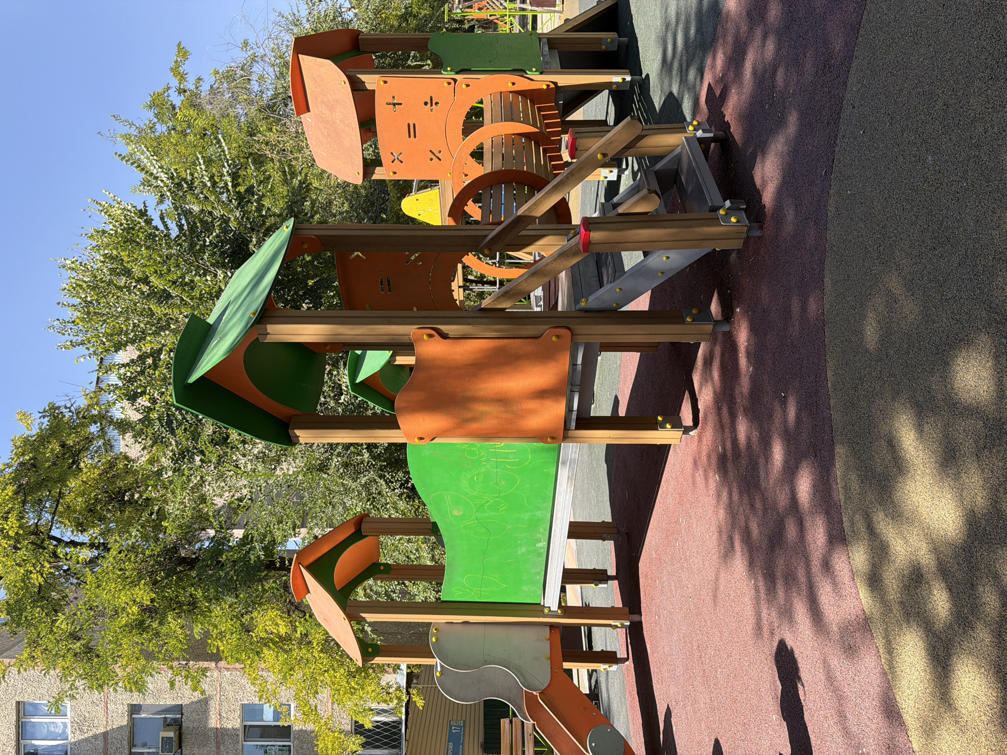 The image shows a children's playground with a multi-level play structure. The structure is made of wood and brightly colored plastic, featuring slides, stairs, and decorative elements resembling train cars. The playground surface is made of red and green rubberized material. In the background, a building with windows and air conditioning units is visible, along with lush green trees and a clear blue sky. The scene is illuminated by bright sunlight, casting shadows on the playground. No people are visible in the image. The location is identified as Shymkent, Kazakhstan, likely in a residential area given the adjacent building.