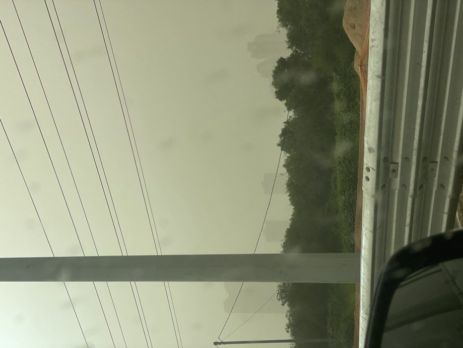 The image depicts an outdoor urban landscape in Ramat HaSharon, Israel, obscured by a dense, yellowish-brown atmospheric haze, characteristic of a dust storm, significantly reducing visibility. The photograph appears to be taken from inside a vehicle, indicated by a partial view of a dark car's side mirror and frame in the lower left, and faint circular smudges on what appears to be a window.
In the foreground, a metallic, multi-section guardrail runs horizontally across the lower portion of the frame, bolted together at regular intervals. Behind the guardrail, light brown earth and an orange construction safety net are visible, along with a small pile of sand to the right.
The midground features a line of dense, dark green trees and shrubs. A tall, light grey utility pole stands vertically in the left-center, extending towards the top of the frame. Multiple dark, thin utility lines stretch horizontally across the hazy sky, seemingly connected to this pole and other, less distinct structures. A smaller, darker utility pole is partially visible further left, carrying at least one wire.
In the background, the silhouettes of several multi-story buildings, consistent with an urban skyline, are faintly discernible through the thick haze. Their forms are indistinct and heavily blurred due to the atmospheric conditions. No individuals or explicit actions are visible within the frame.
