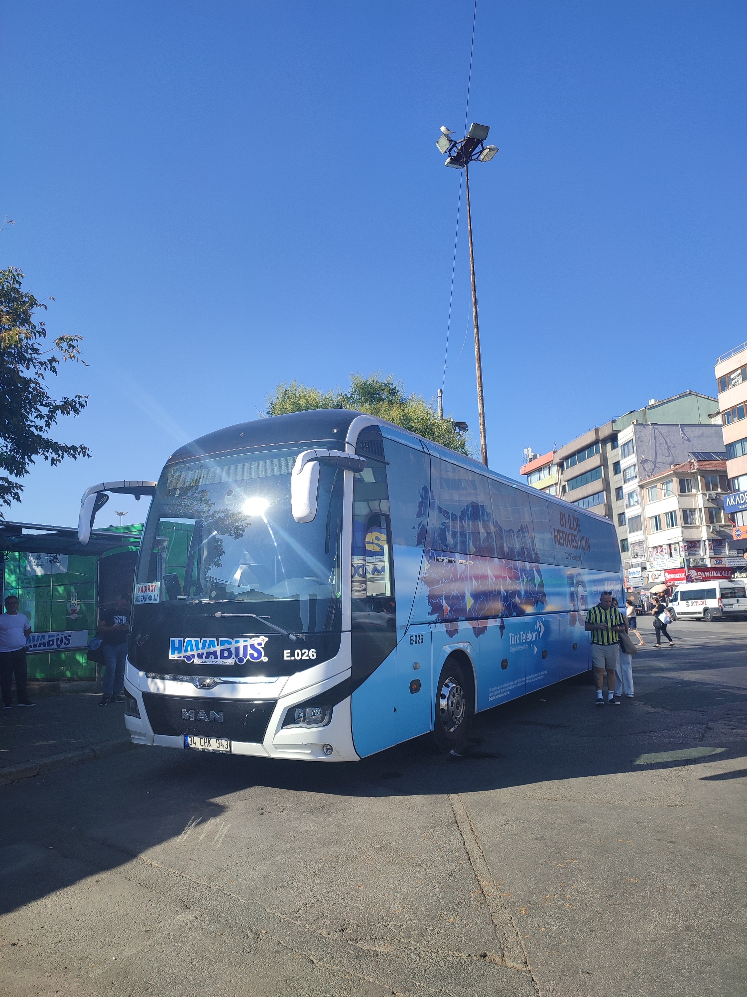 The image depicts a sunny outdoor scene in Kadıköy, Turkey, with a large blue and white "HAVABUS" airport shuttle bus prominently featured in the foreground. The bus, identified by the license plate "34 CHK 943" and model designation "E.026", is parked on an asphalt surface. The bus is decorated with a world map graphic and text that reads "81 İLDE HERKES İÇİN" (For everyone in 81 provinces) and "Türk Telekom" (Turkish Telecom). Several individuals are visible near the bus and in the background. Two men stand on the left, one in a white t-shirt and another in a dark shirt. A man in a striped yellow and green shirt and shorts stands to the right of the bus, looking at his phone. Two women walk in the background to the right, and a white van is also visible on the street. The environment includes buildings in the background, streetlights on a tall pole, and trees on the left side of the frame. The sky is clear blue.