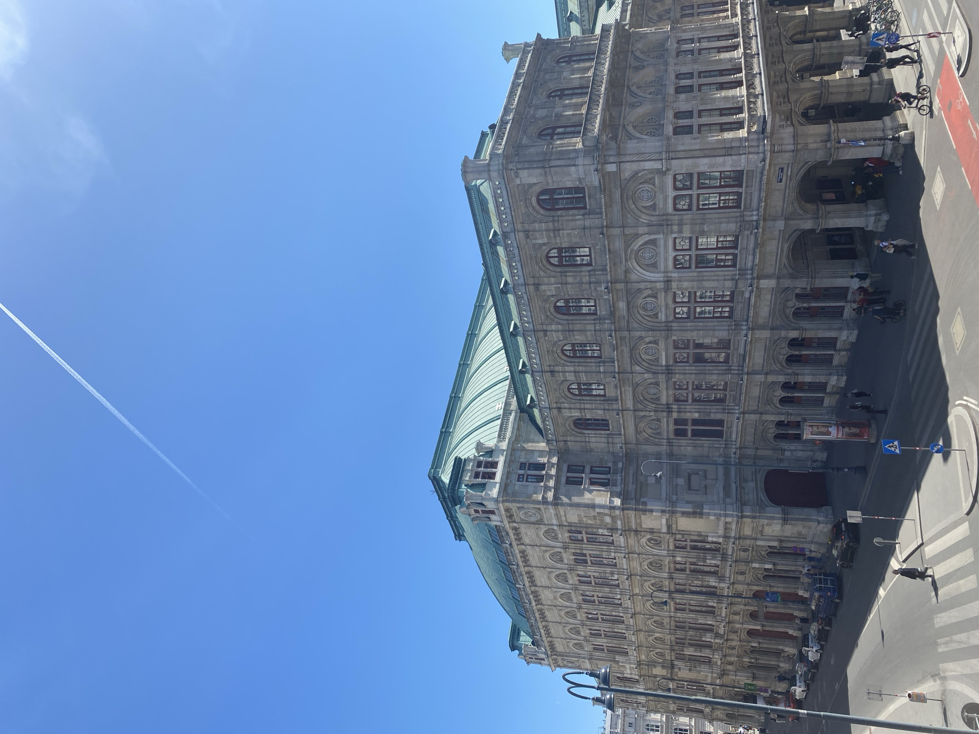 The image shows the Vienna State Opera building in Vienna, Austria. The building is a large, ornate structure with a green roof. There are people walking and cycling on the sidewalk in front of the building. There are cars parked on the street. The sky is blue and clear, and a contrail can be seen.