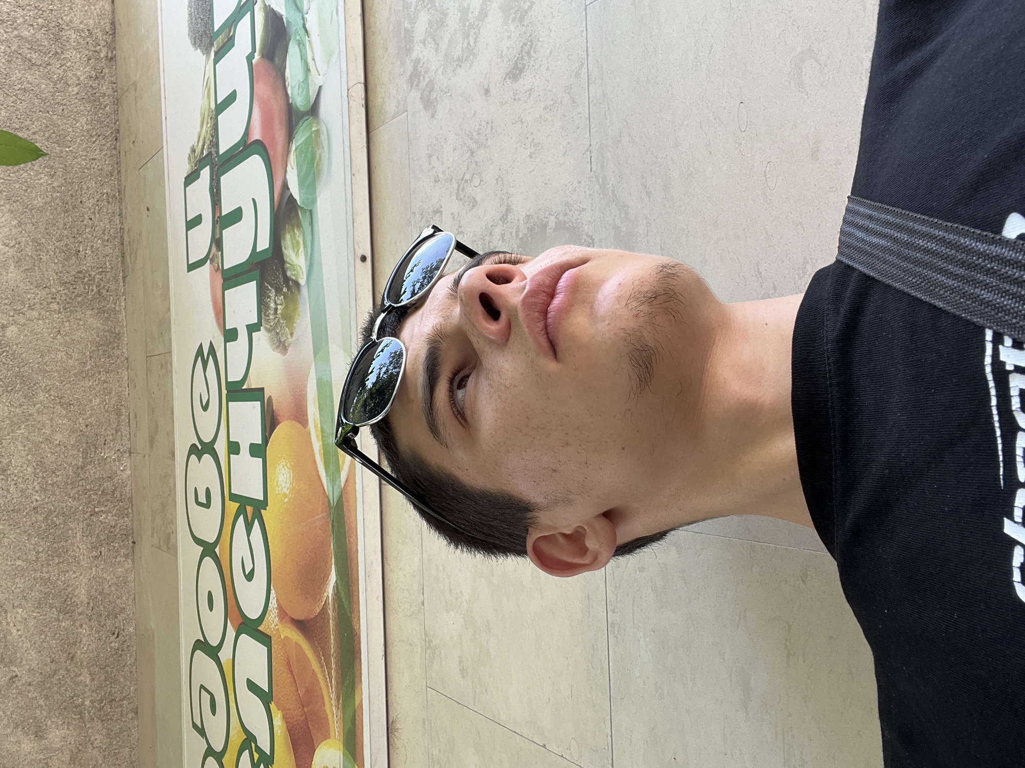 A low-angle shot in Karlovo, Bulgaria, captures a young male looking upwards. He has short, dark hair, minimal facial hair, and sunglasses perched on his head, reflecting the sky. He wears a black t-shirt with a white partial logo visible. A dark grey, textured shoulder strap crosses his right shoulder. Behind and above him, a horizontal sign features white and green Cyrillic text, including "добе" and "счу", overlaying images of sliced oranges and various green vegetables such as lettuce, broccoli, and pickles. Above the sign and to the sides, a textured, light tan wall is visible. A single green leaf hangs into the frame from the upper right corner.