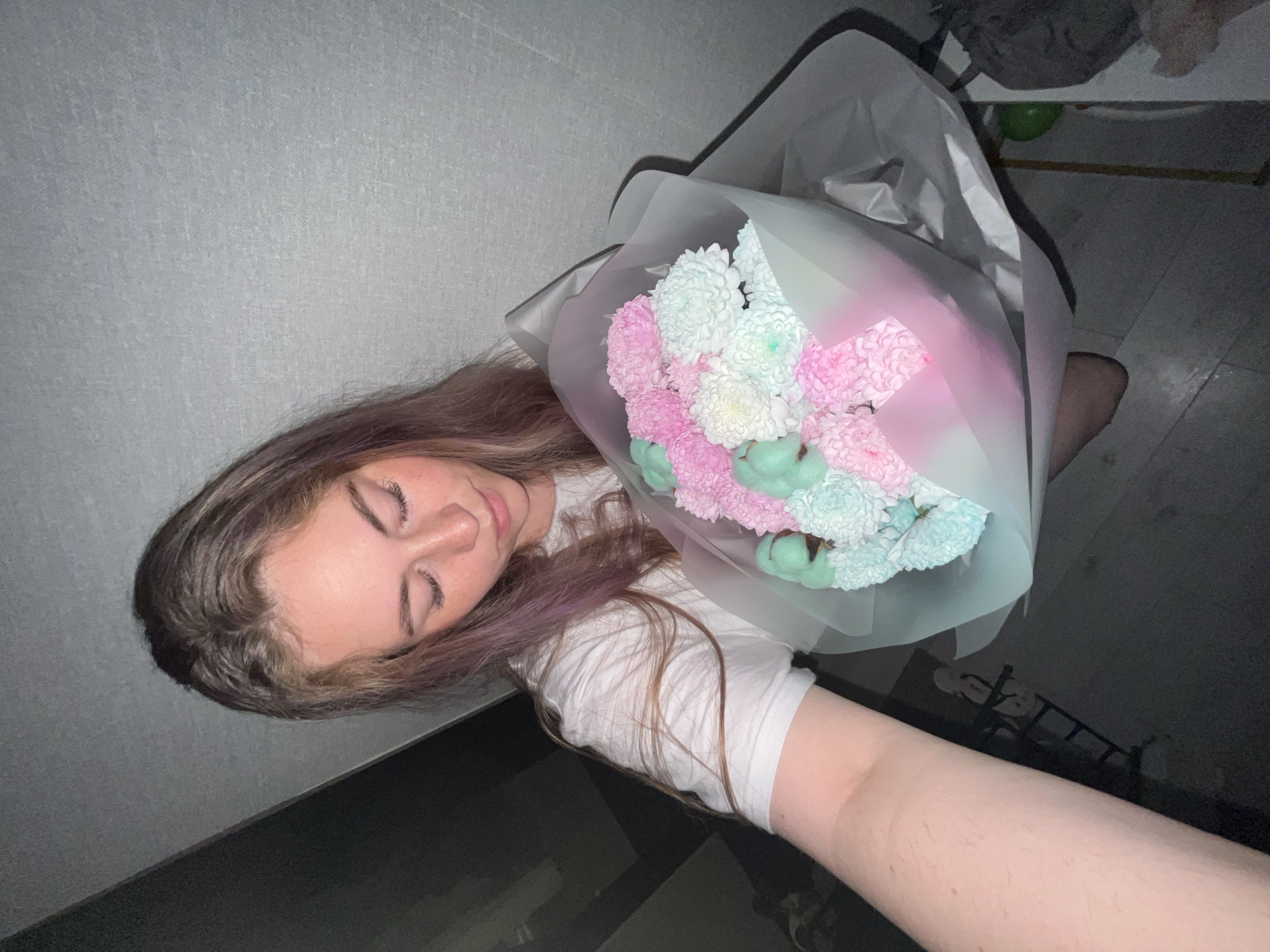 A young person with light skin, brown hair with hints of purple, and closed eyes is holding a bouquet of flowers. The bouquet contains pink, white, and light blue chrysanthemums, and green cotton. The flowers are wrapped in translucent white and light blue packaging. The person is wearing a white t-shirt. The background includes a gray wall and a dark floor. It is not possible to ascertain that this photo was taken in the city of Chaikovsky, Russia.