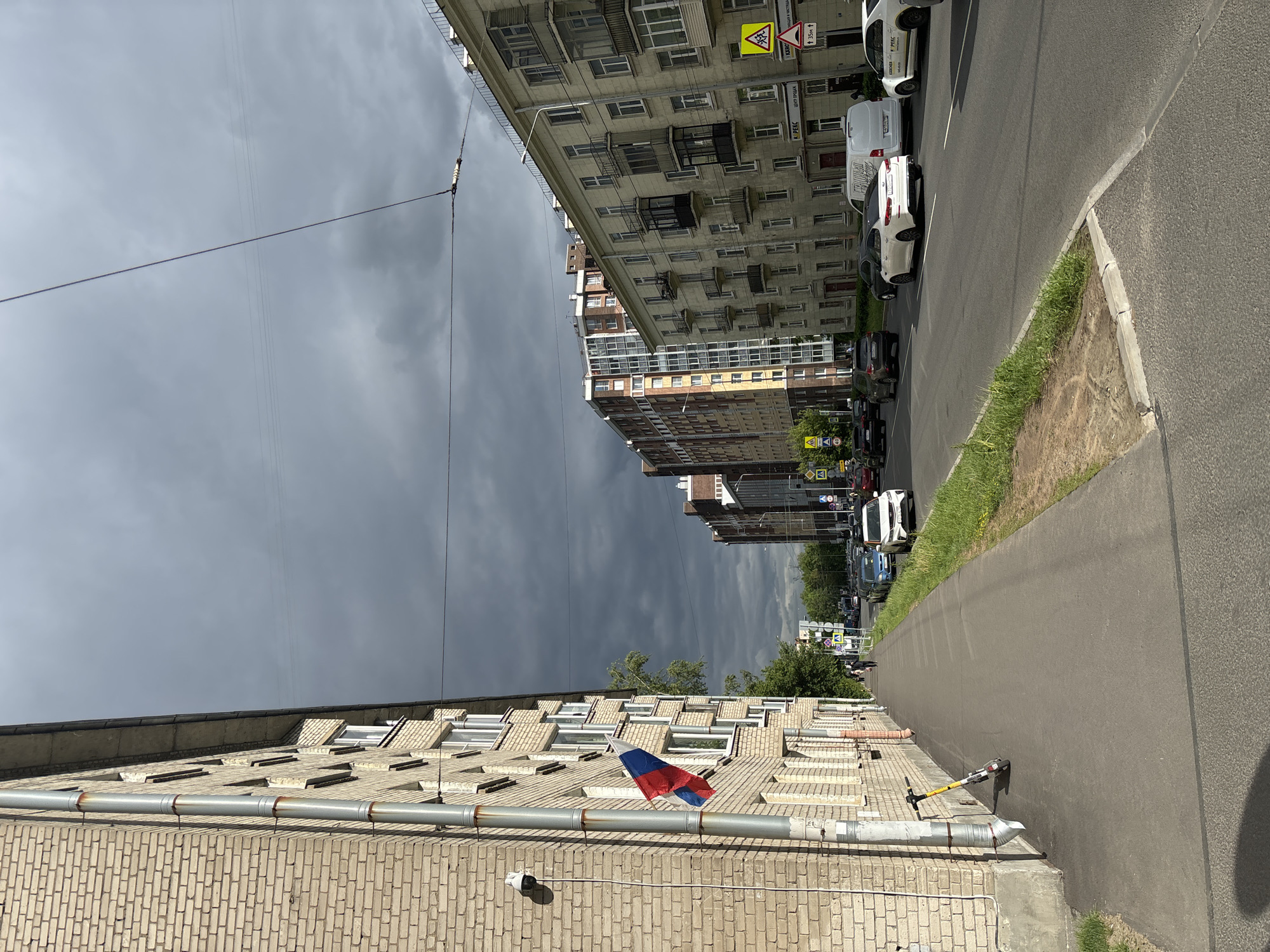 The image depicts a street scene in an urban area with overcast skies. Buildings line both sides of the street. A brick building is visible on the lower left, adorned with a Russian flag. An object resembling a scooter is leaned against the building's wall. Cars are parked along the right side of the street. Traffic signs and other buildings are visible further down the street, indicating a typical city environment.