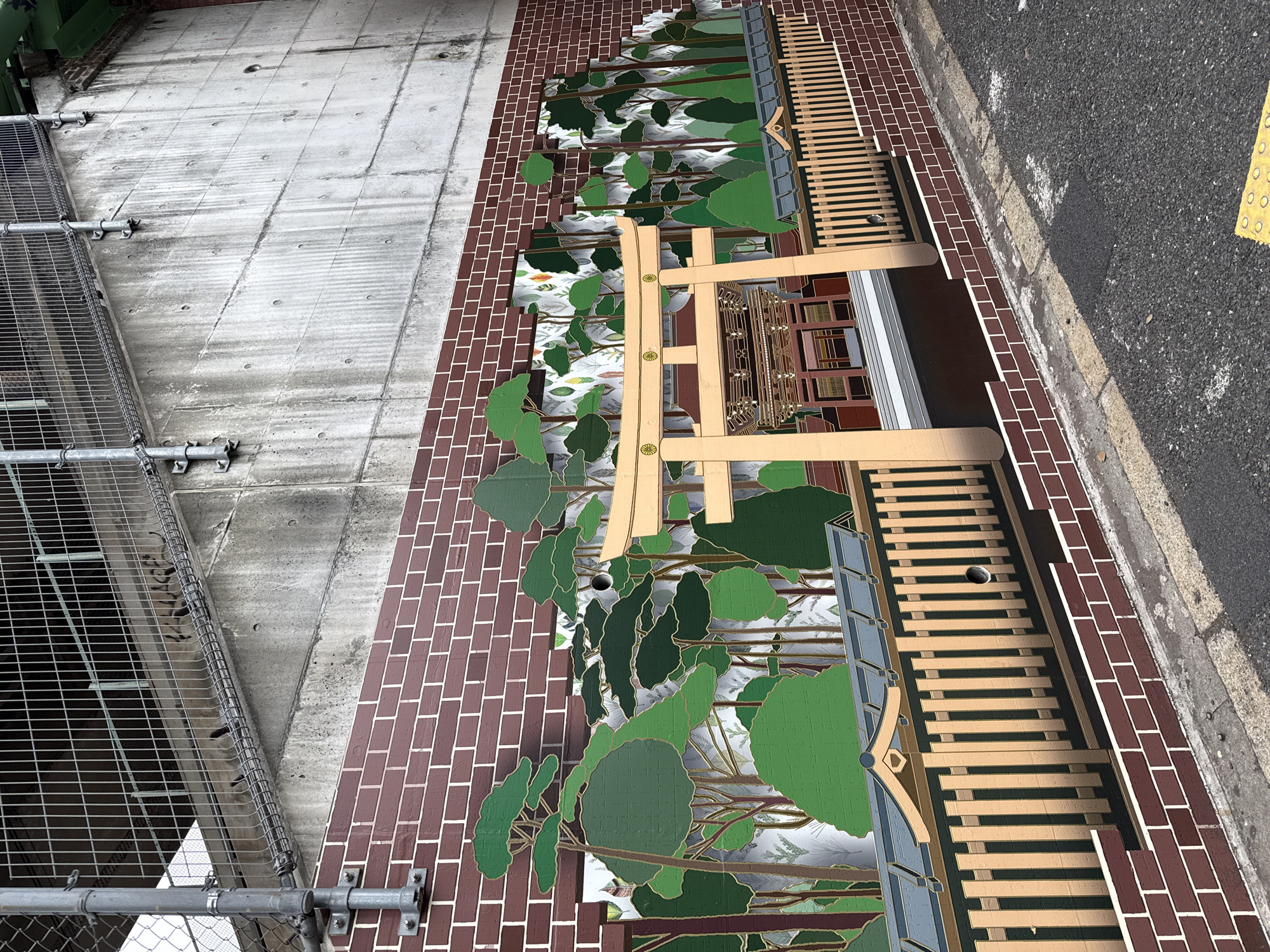 This image presents an outdoor urban scene, likely viewed from an elevated perspective, showcasing a blend of industrial architecture, public art, and street infrastructure.
On the left, a large, textured concrete wall or support structure is visible. It's grey, marked with rectangular divisions and signs of weathering, and features several metal bolts and fixtures. A robust metal scaffolding or fence structure is attached to the far left side of this concrete surface.
Adjacent to the concrete, a prominent vertical surface decorated with a deep red brick pattern dominates the central area. Adorning this brickwork is a large, stylized mural depicting a dense forest scene. The mural features numerous trees and foliage in various shades of green, outlined in gold, against a lighter, soft grey-blue background, creating a layered and somewhat whimsical aesthetic.
Integrated with or in front of this mural is a light-colored wooden structure, resembling a traditional Japanese torii gate, with its distinctive crossbeams and uprights. Below the torii gate, part of the mural depicts a building facade with windows and doors, suggesting a shop or house. Further right along the brick-patterned wall, another wooden structure, composed of horizontal slats, extends, possibly acting as a decorative fence or seating.
The far right of the image reveals a stretch of dark asphalt road, bordered by a concrete curb and a yellow tactile paving strip, commonly used for the visually impaired in Japan. The overall lighting suggests daytime, likely overcast, as there are no harsh shadows. No people or visible text are present, and the scene depicts a static urban landscape featuring decorative public art. The architectural and street elements are consistent with an urban setting in Shibuya, Japan.