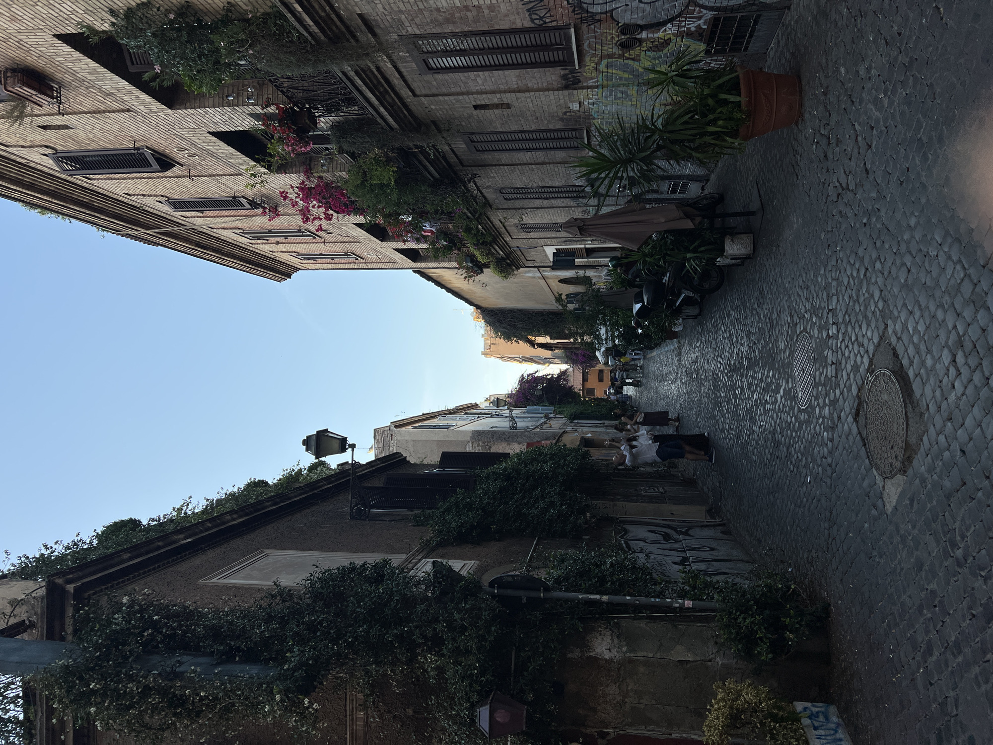 The image depicts a narrow, historic cobblestone street or alleyway, characteristic of an old European city like Rome, under a clear blue sky during the daytime. The street is lined on both sides by multi-story buildings, featuring facades of brick and stucco with numerous windows often fitted with dark wooden or metal shutters.
Abundant greenery decorates the scene, with ivy climbing walls, lush plants in window boxes and on balconies, and vibrant pink flowering plants (resembling bougainvillea) adding splashes of color. Large potted plants, including a prominent yucca-like plant in a terracotta pot, are placed along the street. Traditional lampposts are visible, providing ambient lighting.
The cobblestone pavement is well-maintained and features several circular manhole covers. On the right side of the street, a few tables and chairs are set up under a partially open brown umbrella, indicating an outdoor dining area or cafe. Several motor scooters or mopeds are parked alongside the building on the right.
A few people can be seen further down the street on the right, some seated and others standing casually, suggesting everyday activity. The overall atmosphere appears calm and pleasant. Graffiti is visible on the upper section of the right-hand building, with parts of the word "GRAF" discernible among other markings.