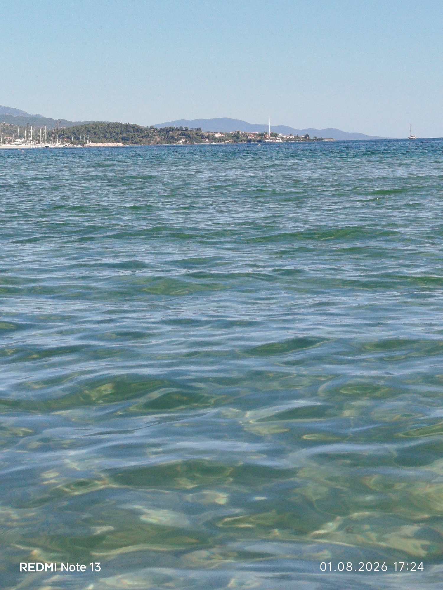 The image displays a tranquil sea scene under a clear blue sky. In the distance, a coastline is visible, characterized by a forested hill dotted with buildings and a marina filled with numerous sailboats. The foreground is dominated by the rippling surface of the turquoise water, revealing hints of the sandy seabed beneath. The overall impression is one of a peaceful summer day at a coastal location. Based on the context of the surrounding text overlay which mentions "Sithonia Municipal Unit, Greece", the location is likely within this region, known for its scenic beauty and numerous coastal settlements.