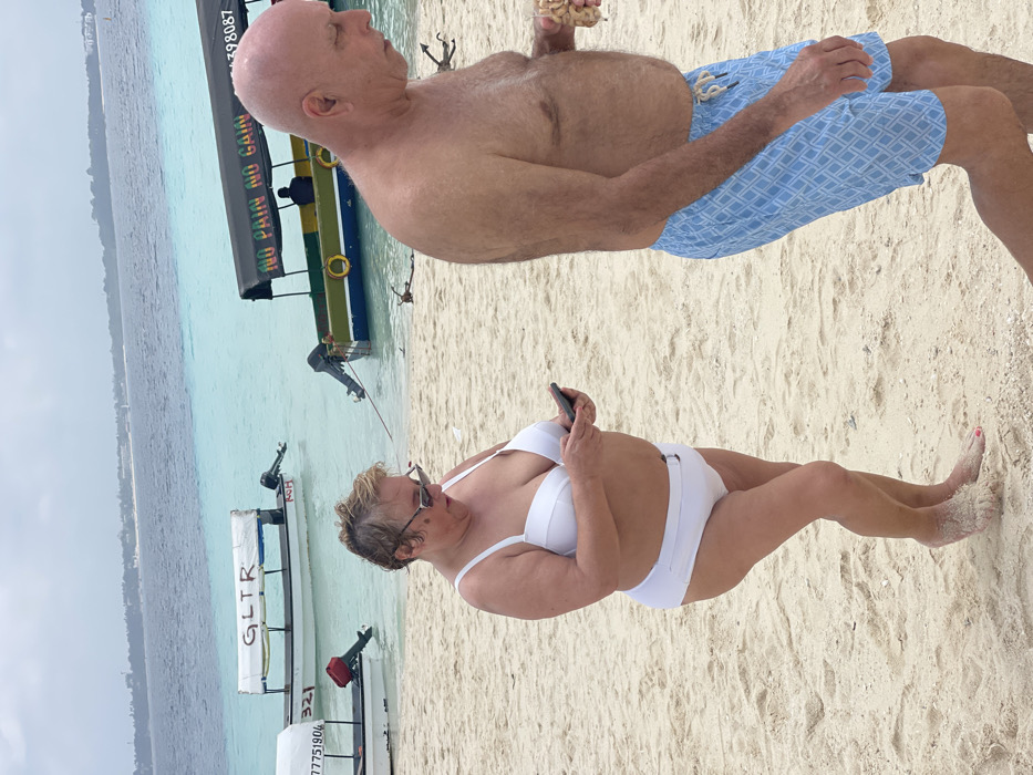 The scene depicts a beach in Zanzibar, Tanzania, under an overcast sky. In the foreground, a middle-aged woman with short, light-colored hair and sunglasses stands barefoot on light-colored sand, angled slightly right. She wears a white two-piece swimsuit and is engaged with a black smartphone held in both hands, looking down at its screen. Sand adheres to her feet; her left big toenail is painted pink. To her right and slightly behind, a bald, shirtless middle-aged man walks towards the right side of the frame. He wears light blue swim trunks with a white geometric pattern and carries a clear plastic cup filled with light-colored snacks in his right hand. He looks downwards.
The background features several small, motorized boats moored in shallow, clear turquoise water, which transitions to a deeper blue further from shore. One boat displays "GLTR" on its white canopy, another bears "77751904" and "321" on its hull, and a third, painted green, yellow, and blue, has "NO PAIN NO GAIN" written on its dark canopy, with two orange life preservers attached to its side. A faint landmass with visible structures is present on the distant horizon. Minimal natural debris is scattered in the water and on the immediate shoreline.