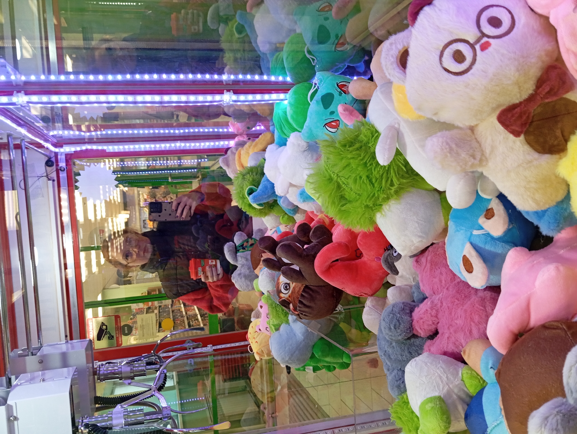 A claw machine filled with plush toys, including characters resembling Bulbasaur and other various creatures, is the primary focus of the image. The machine's interior is illuminated with LED strips. The reflection in the machine's glass shows a person holding a smartphone and a small orange container, seemingly taking a picture. The person has short, curly hair, is wearing glasses, and is wearing a black jacket over a red shirt. Behind the person's reflection are posters and merchandise displays. The location is Yurgamysh, Russia.