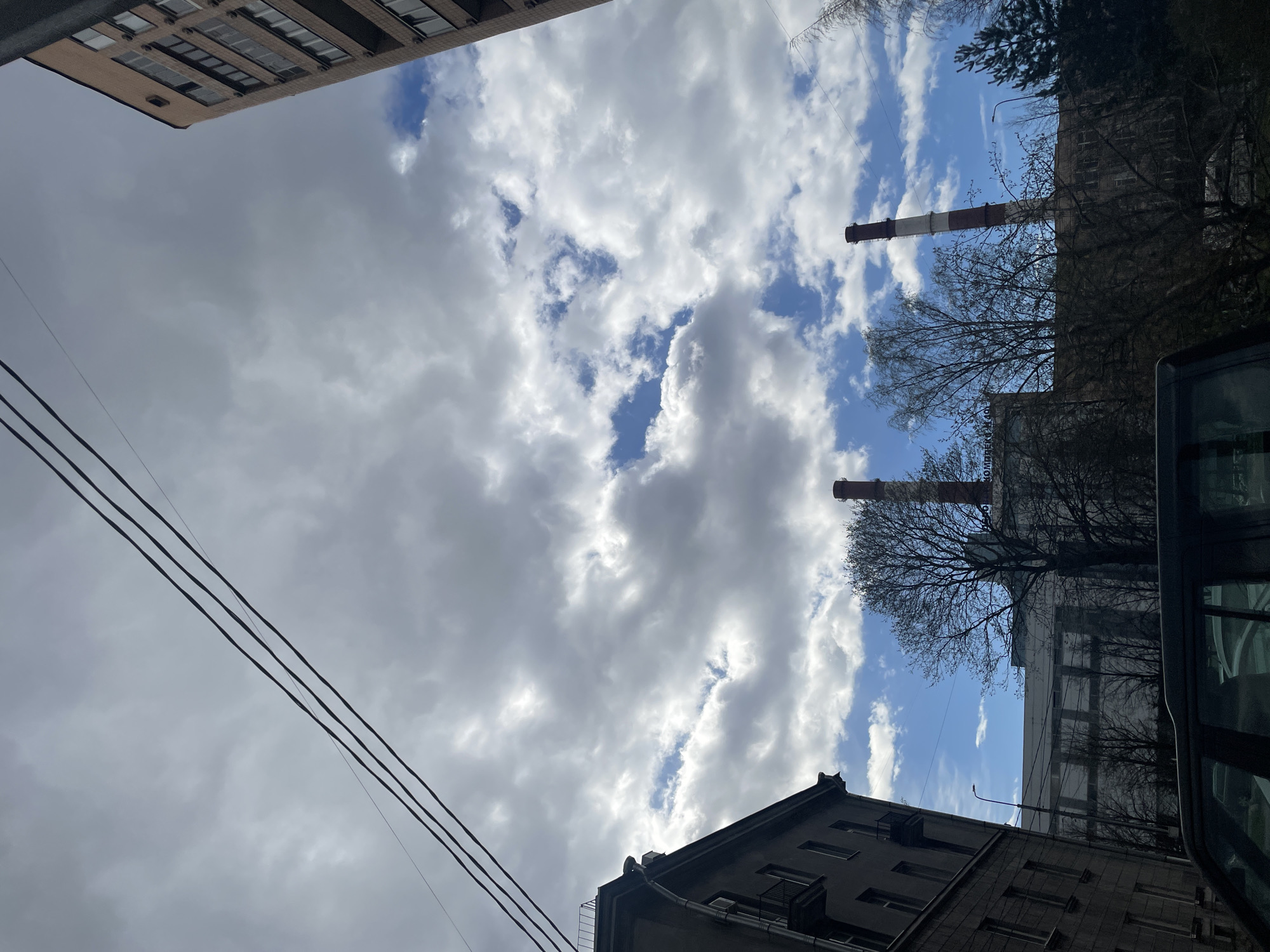 An upward-angled outdoor shot captures a dynamic sky above an urban or industrial landscape in Vsevolozhsk, Russia. The sky is a prominent feature, displaying a mix of bright blue patches and large, fluffy white-grey clouds, suggesting a partly cloudy daytime.
The scene includes elements of urban infrastructure and architecture:
- Parts of several buildings are visible from a low vantage point: a light-colored multi-story building with numerous windows occupies the top-left, while a darker-toned building with windows and an air conditioning unit is seen from its corner in the bottom-right. Further back on the right, a larger, light-colored industrial-style building is partially obscured by trees.
- Two tall, slender industrial chimneys, both striped in red and white, stand against the sky on the right side of the frame.
- Numerous bare, leafless trees are visible on the right, indicating a cooler season like late autumn, winter, or early spring.
- Three dark power lines or utility cables stretch diagonally across the mid-left portion of the image.
- A streetlight is faintly visible through the trees on the right.
- The dark frame and window of a vehicle are visible on the far right, suggesting the photo was taken from within or immediately beside it.
Faint Cyrillic text, possibly "ОМВЕЛКРО" or similar, can be discerned on the industrial building behind one of the chimneys. The overall impression is one of a typical industrial or residential area under a vast sky.