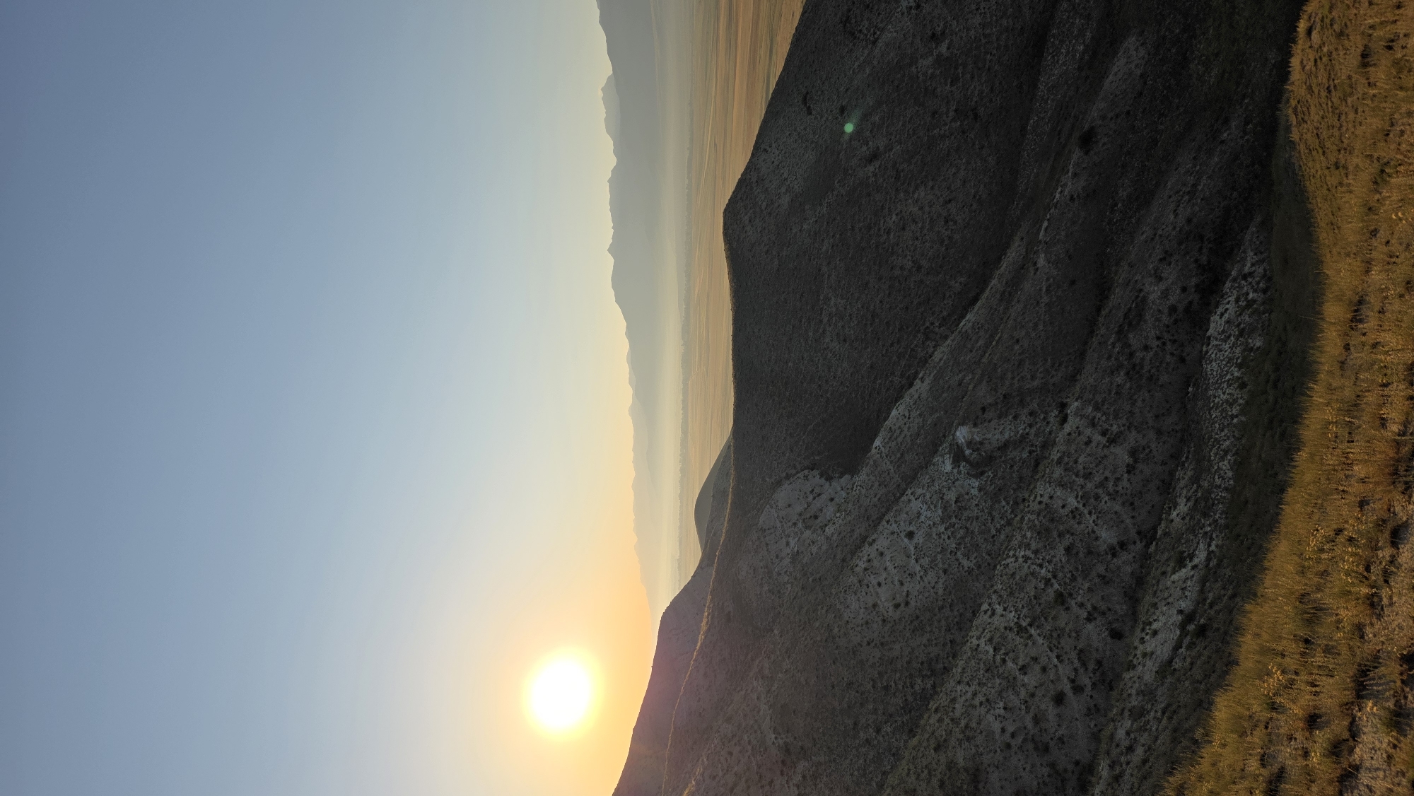 An outdoor, vertical, full shot captures a sunrise over a vast valley and mountain range. The sun, a bright orb, is positioned on the left side of the frame, casting a warm glow across the sky, which transitions from a pale yellow near the horizon to a clear, light blue at the top. In the foreground, rolling hills covered in sparse vegetation and rocks dominate the scene. The hills are cast in shadow, with the rising sun illuminating their upper slopes and the grass at the very bottom of the frame. Beyond the foreground hills, a wide, flat valley stretches into the distance. The valley appears to be agricultural land, with geometric patterns of fields visible. A light haze or fog blankets the valley floor and the base of the distant mountain range, softening the landscape. The mountains themselves are a series of silhouetted peaks, layered against the horizon, with a lighter, more ethereal appearance due to the distance and atmospheric conditions. The scene is tranquil and serene, evoking a sense of natural beauty and vastness. The provided metadata does not contain information about the location of Dyikan, Kyrgyzstan.