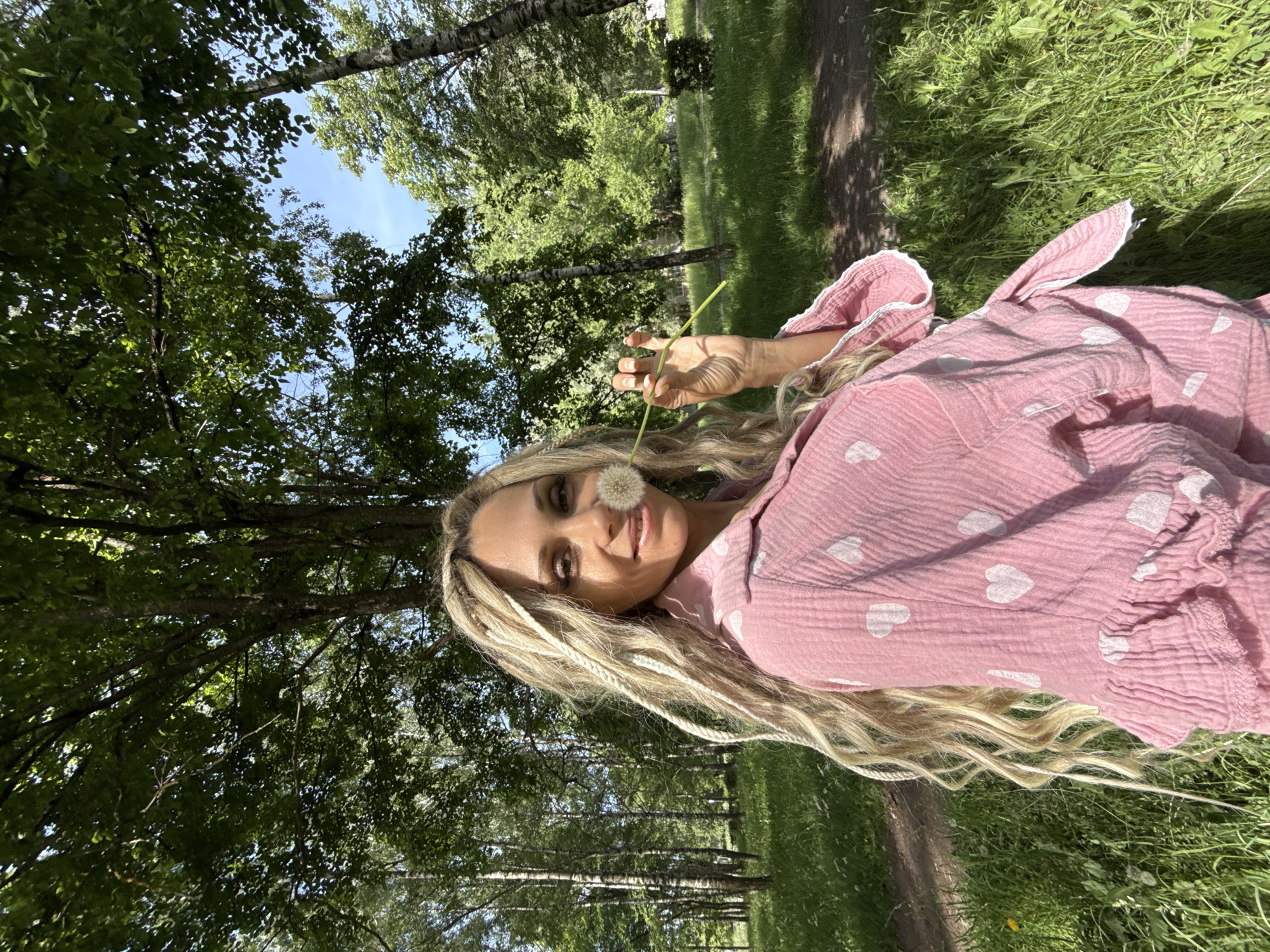 The image depicts a woman standing in a sunlit park or wooded area. She is positioned slightly to the left of the frame, facing forward with a slight smile. Her hair is long, blonde, and wavy, featuring several interwoven blonde dreadlock-style braids. She is wearing a pink, textured shirt or top adorned with white heart patterns. The garment has a crinkled appearance and a ruffled hem or cuff. In her right hand, she holds a single dandelion puffball on its stem, positioned near her nose and mouth. The background consists of numerous deciduous trees with dense green foliage, suggesting a lush, outdoor environment. Patches of blue sky are visible through the tree canopy. The ground is covered with green grass and visible dirt paths. The scene is brightly lit by natural sunlight, creating varied shadows and highlights.