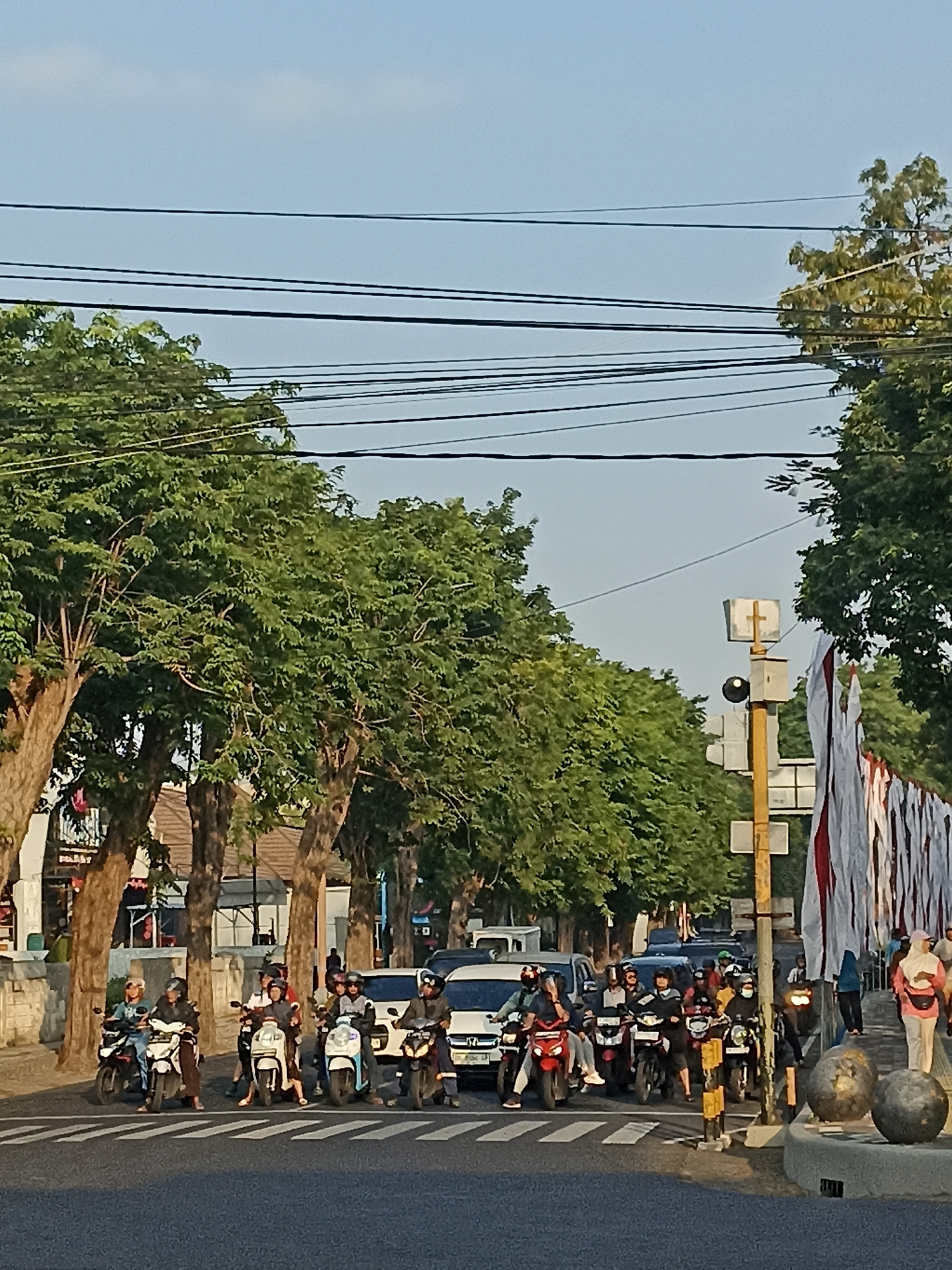 The image shows a street scene in Kelurahan Kepatihan, Indonesia, with a line of motorcycles and cars at a crosswalk. Several people are riding on the motorcycles, most of whom are wearing helmets. There are trees lining the street, and a few buildings can be seen in the background. A traffic light and several flags are also visible. The sky is clear and blue. The overall impression is a busy urban street with moderate traffic.