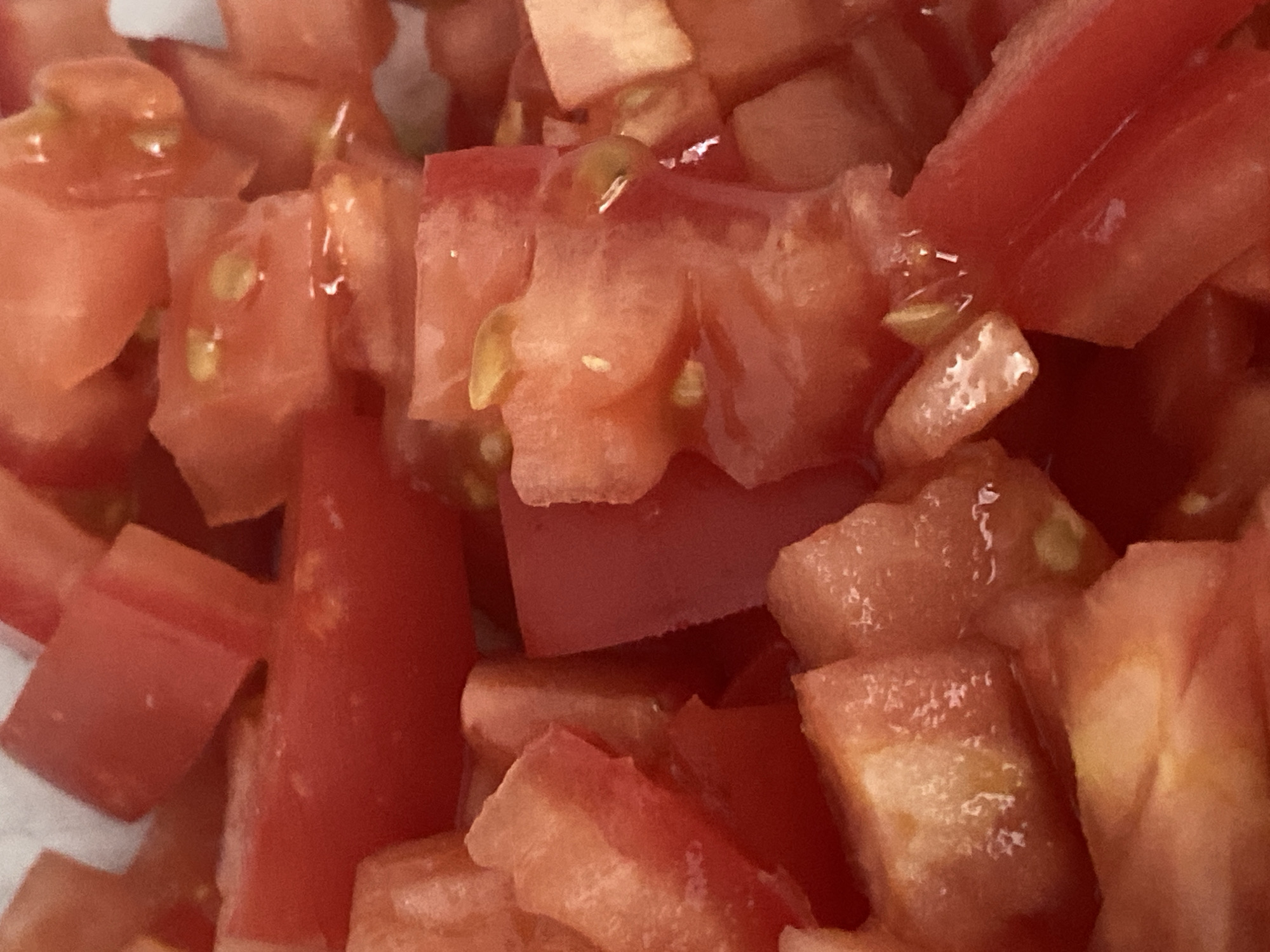 This is a close-up, full-frame photograph of diced tomatoes. The tomatoes are bright red with some lighter pinkish-red hues. The diced pieces vary in size and shape, with some being roughly cubic and others more irregular. Small, pale yellow seeds are visible within some of the tomato pieces, along with some of the juicy pulp. The lighting appears to be overhead and slightly diffused, creating subtle highlights on the moist surfaces of the tomatoes. There is no discernible background or environment visible, as the image is entirely filled with the tomato pieces. The location in the city of Balashikha, Russia is not discernible from the image.