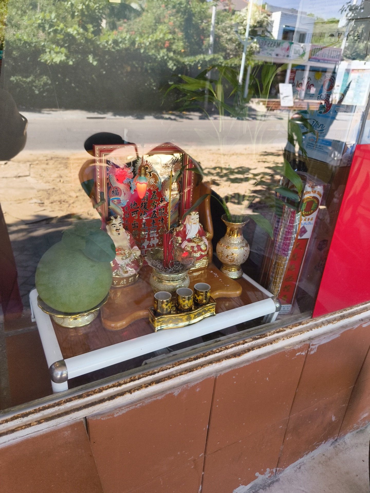 The image shows a shrine inside a shop window in Phu Thuy Ward, Vietnam. The shrine features various items, including a large green fruit, a golden vase, two figurines of elderly men in red robes, and an incense burner with sticks. Behind the figurines is a framed picture with a red dragon and text. Several small golden cups are arranged on a tray. The shrine is placed on a white table with a wooden top. The background outside the window shows a road, trees, and buildings, suggesting an urban environment. The glass reflects the surroundings, including trees and a partial view of a building with signage. The foreground shows the lower part of the shop's exterior, with reddish-brown tiles and a white frame.