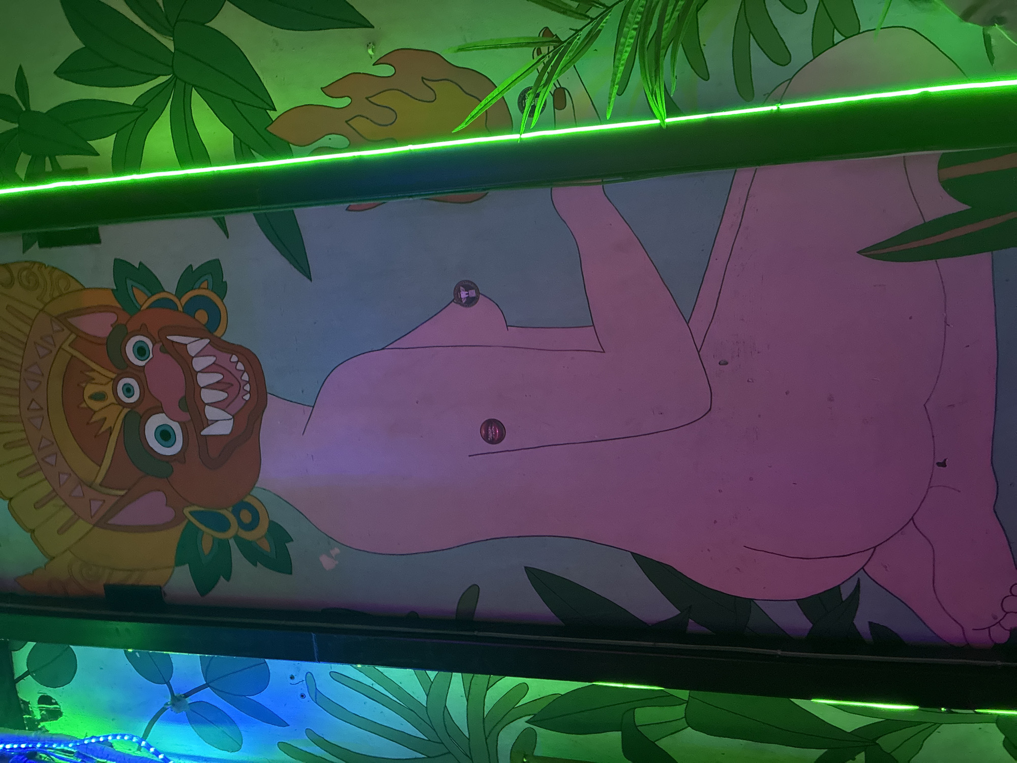 A mural depicting a three-eyed anthropomorphic figure with exposed breasts is visible. The figure's head resembles a feline or mythological creature with sharp teeth and a golden headpiece. The body is depicted in a reclined position, and the scene is framed by stylized tropical foliage. The mural is lit by a green neon light. The location is Tel-Aviv, Israel.
