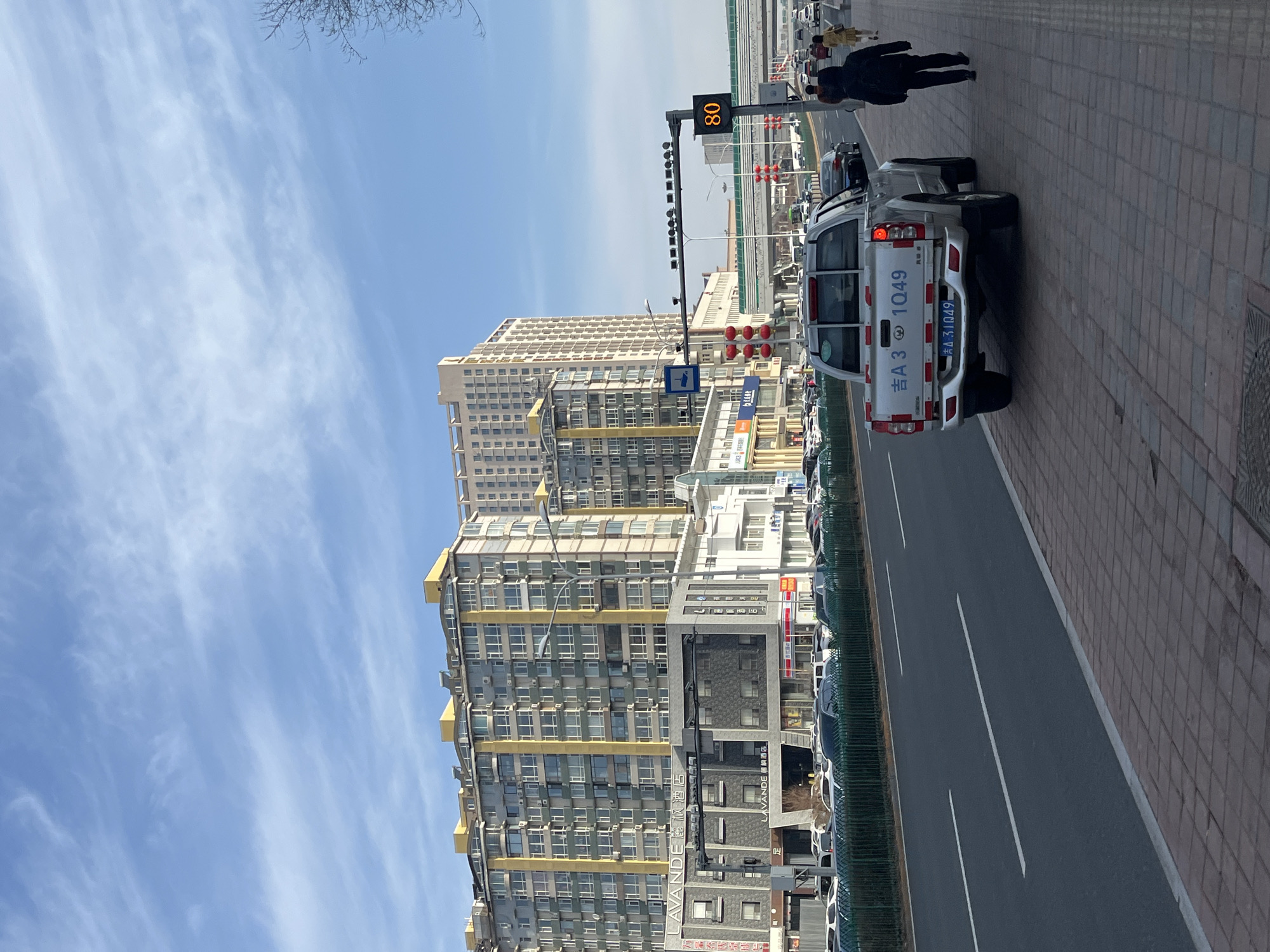 The image captures a street scene in Changchun City, China. A white pickup truck with the license plate "吉A 3 1Q49" is parked on the brick-paved sidewalk adjacent to the asphalt road, which has lane markings. A digital speed limit sign displaying "80" stands near the road. Pedestrians are walking on the sidewalk. The backdrop features a building complex with grey and yellow accents, with "LAVANDE 麗楓酒店" signage visible on one of the buildings. In the distance is a multi-story apartment building under a blue sky with streaks of clouds.
