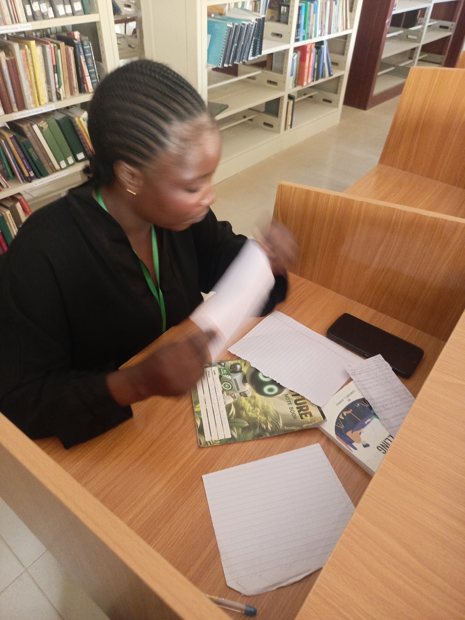 A woman is sitting at a desk in a library in Akwanga, Nigeria. She has braided hair, dark skin, and is wearing a black shirt with a green lanyard. She is holding a piece of paper in her hands. On the desk are two notebooks, some loose sheets of paper, a pen, and a black smartphone. The desk and chair are made of light-colored wood. Bookshelves are visible in the background, filled with books.