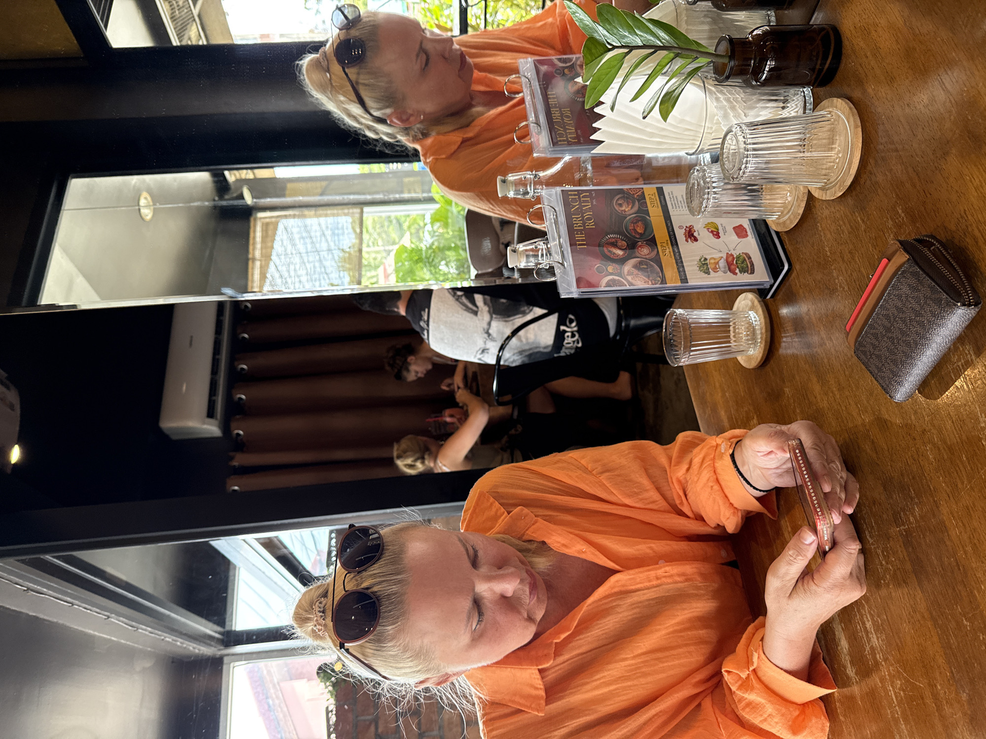 The image depicts a scene inside a restaurant in Ko Samui District, Thailand. Two women are seated at a wooden table. The woman in the foreground, facing left, is wearing an orange blouse and looking down at her phone, which she holds in her hands. She has blonde hair pulled up in a bun with sunglasses perched on top. To her right, another woman, also with blonde hair, is visible from the side, wearing a similar orange top and looking down at a menu on the table. Several items are on the table, including a wallet, two empty glasses, a menu, a bottle of water, and a small vase with a plant. In the background, other patrons can be seen, and reflections in a mirror reveal more of the restaurant's interior. The environment appears to be brightly lit, with natural light coming through windows and artificial lighting from above.
