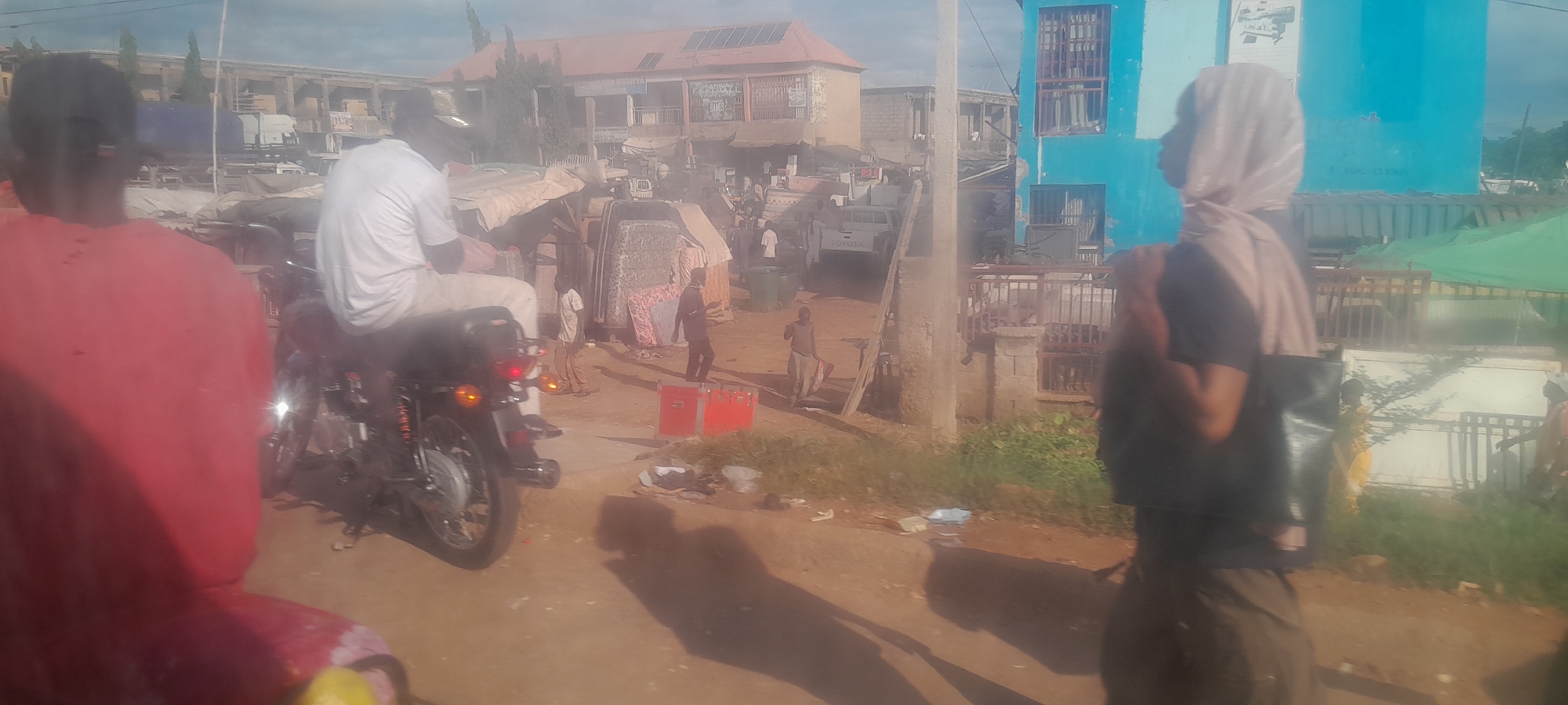 In Nyanya, Nigeria, a street scene unfolds with a mix of residential and commercial activity. In the foreground, a person in a red garment faces away from the camera, their presence casting a prominent shadow. To their left, a motorcycle is parked, with a person in a white t-shirt seated on it, also facing away. Further into the scene, a building with a reddish-orange roof and solar panels stands against a hazy sky. Adjacent to it, a bright blue building with multiple windows is visible. The street is dusty, with scattered debris. Several individuals are seen walking or standing, engaged in various activities. Some are near stalls or market areas, indicated by the presence of what appear to be mattresses stacked and covered. Red containers are placed on the ground, and a white Toyota pickup truck is parked in the middle ground. A person in the right foreground, wearing a headscarf and dark clothing, is also seen from the side, carrying a bag. The environment suggests a bustling urban setting with a blend of construction, trade, and everyday life.