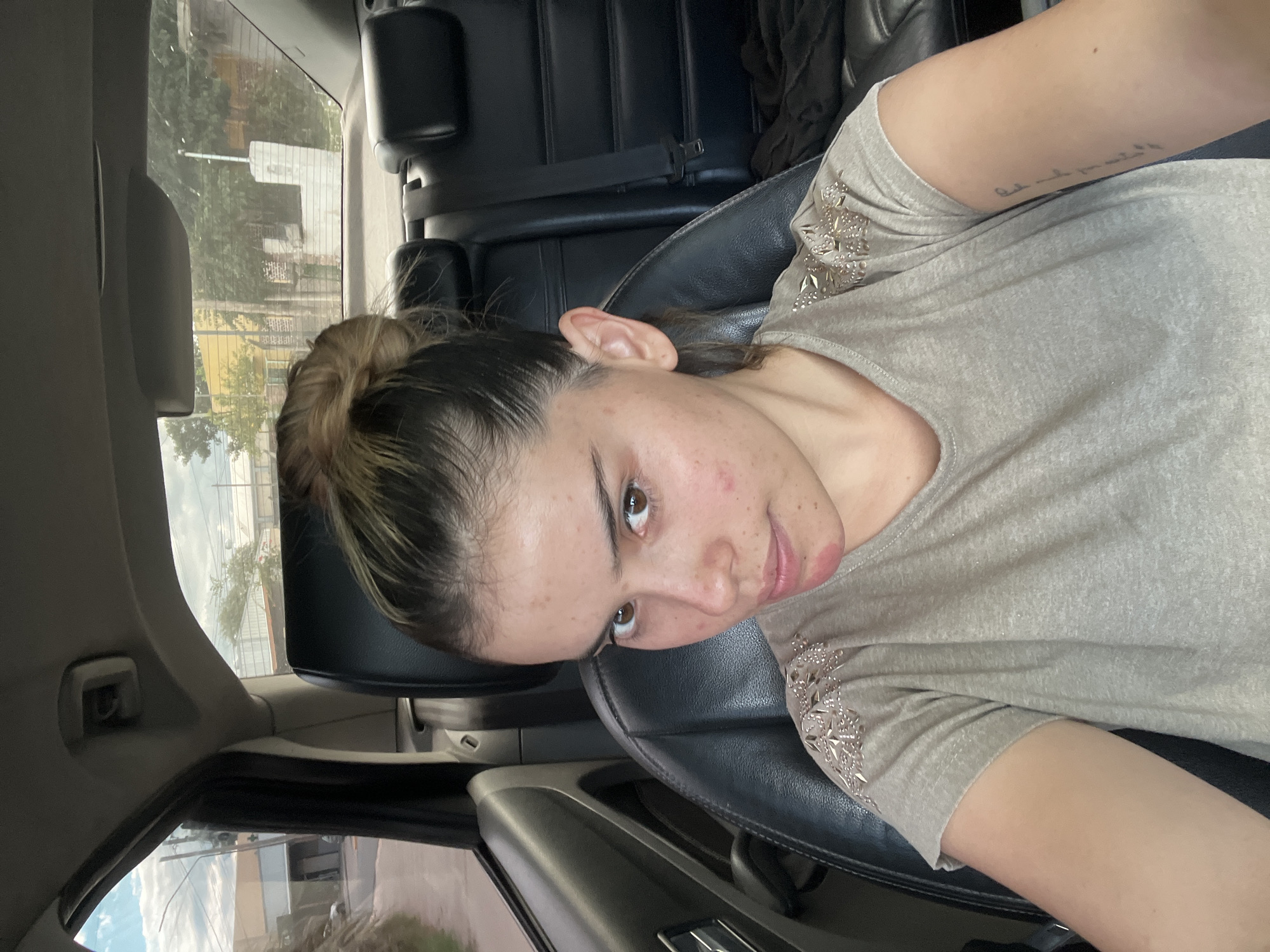 A woman with brown hair pulled back in a bun, wearing a grey shirt with silver embellishments on the sleeves, sits in the driver's seat of a car. The car has black leather seats and interior. Outside the window, the environment appears to be a street or residential area in Sumalao, Argentina, discernible by the architectural styles and vegetation. The image is taken from a slightly high angle, looking down at the woman. She is looking slightly upwards at the camera. A tattoo is visible on her left arm.
