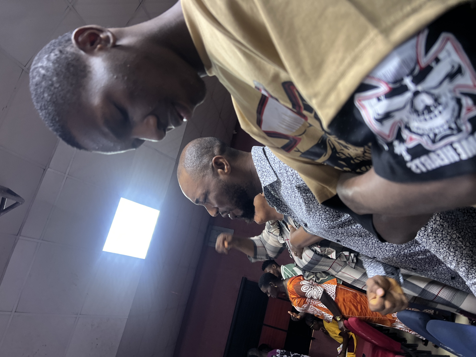 The image depicts an indoor scene, possibly a place of worship or a meeting hall, located in Nkpor, Nigeria. In the foreground, a young Black man is seen in profile, his head bowed and eyes closed. He is wearing a tan t-shirt with a graphic print on it. To his left, an adult Black man with a shaved head and a beard is visible in profile, facing forward with his mouth slightly open, as if speaking or singing. He is wearing a patterned shirt. Behind him, several other individuals are present, including a Black woman wearing a vibrant orange and white patterned dress. The lighting suggests an artificial light source, likely a ceiling fixture, as indicated by the bright square light panel in the upper portion of the image. The walls appear to be a dark reddish-brown color. The overall atmosphere suggests a gathering or a service.