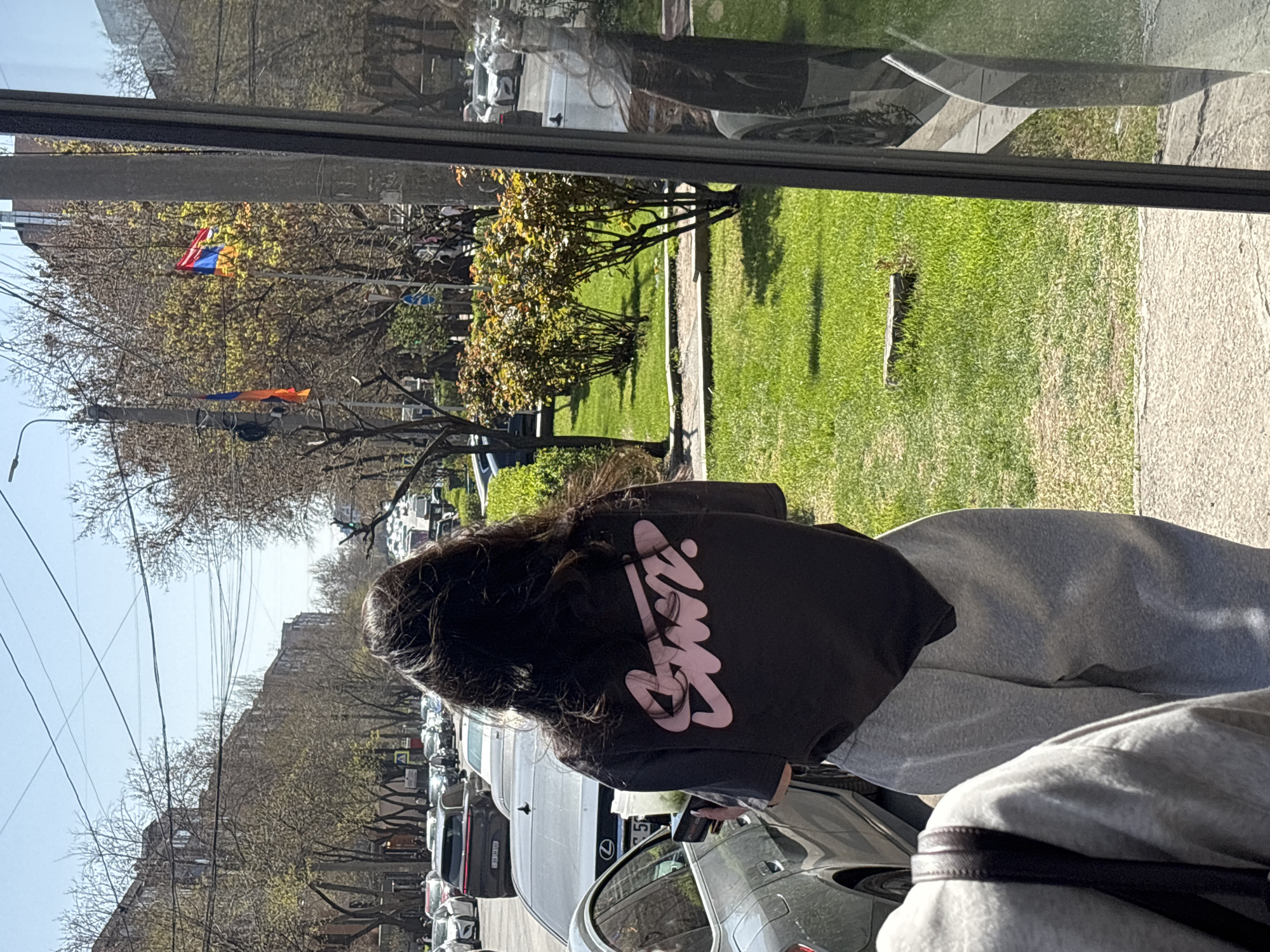 The image captures an outdoor street scene in Yerevan, Armenia, on a bright and sunny day. In the foreground, viewed from behind, is a person with long, dark hair. They are wearing a black long-sleeved top featuring a prominent pink logo that reads "Stussy" in a stylized script, paired with light grey sweatpants. A dark strap from a bag is visible over their right shoulder.
To the left of the person, a street is visible, lined with numerous parked cars. Further in the background, rows of bare-branched trees, interspersed with some showing green leaves, line the street. Beyond these trees, several urban buildings are visible under a clear, pale blue sky, with utility wires crisscrossing overhead.
To the right of the person, a well-maintained grassy area with small bushes and a sidewalk extends. In the mid-ground on this side, two flags are visible flying from poles; the upper flag distinctly displays the red, blue, and orange horizontal stripes of the Armenian national flag, confirming the location.
The upper-right portion of the image shows a large reflective surface, likely a window, which mirrors parts of the street, parked vehicles, and surrounding environment in a distorted manner. The overall scene depicts a typical urban setting during daylight hours, possibly in spring given the mix of bare and budding trees.