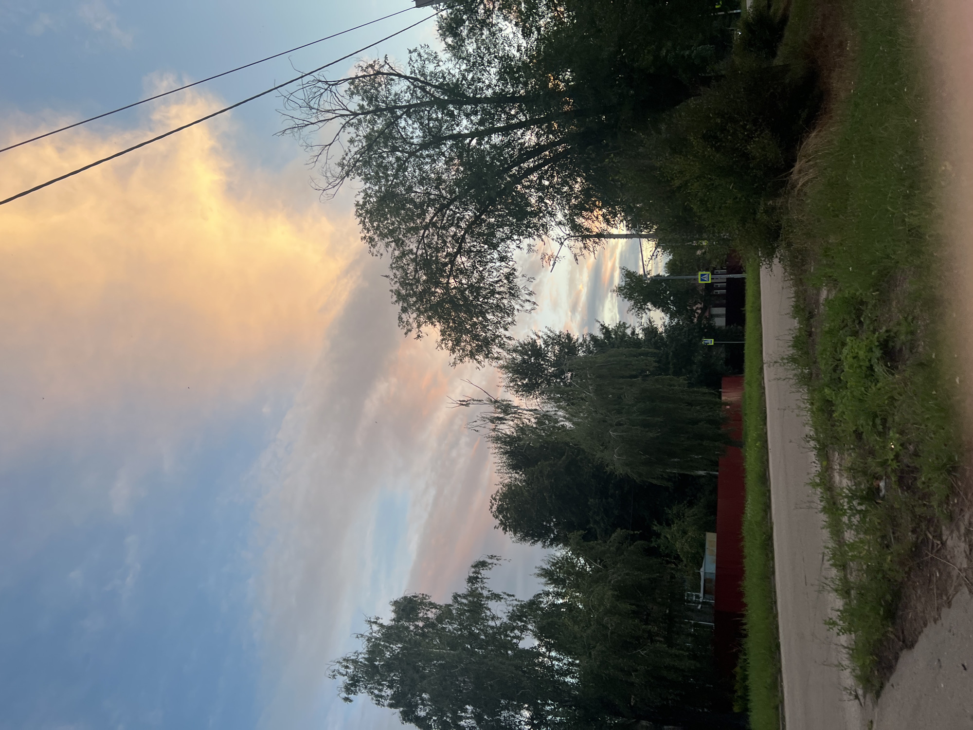 The image shows a rural roadside scene in Ulyanovsk, Russia, during sunset. A paved road runs through the middle of the frame, with a grassy verge on either side. In the background, a red corrugated metal fence encloses a property with some structures. Lush green trees line the roadside, with some appearing to be weeping willows. Above the trees, the sky is a mixture of blue and pastel hues, with wispy clouds catching the last rays of the setting sun, creating a warm, orange glow. Power lines are visible stretching across the upper right portion of the image. A pedestrian crossing sign is visible on the left side of the road. The overall impression is one of a tranquil, semi-rural environment at dusk.