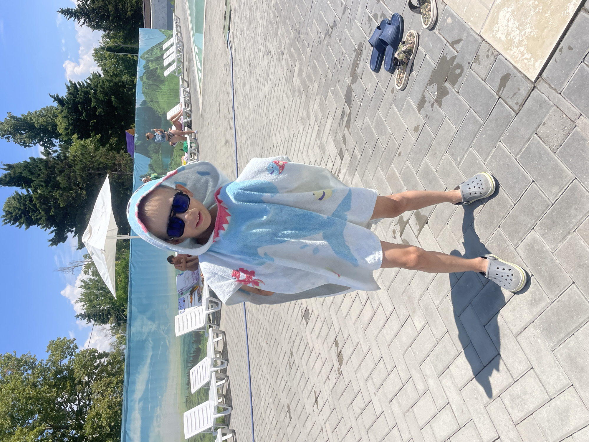 A young child wearing a shark-themed hooded towel and dark sunglasses stands on a paved walkway near a swimming pool in the city of Gagra, Abkhazia. The child is facing the camera, with their body angled slightly to the right. Several white lounge chairs are visible in the background, along with a decorative blue tarp depicting a landscape. In the distance, other people can be seen relaxing by the pool. The ground is made of gray interlocking paving stones, with some damp spots and shadows cast by the sun. A pair of dark blue slides and two camouflage-patterned slides are placed on the ground to the right of the child. The sky is bright blue with scattered white clouds, and lush green trees line the perimeter of the pool area.