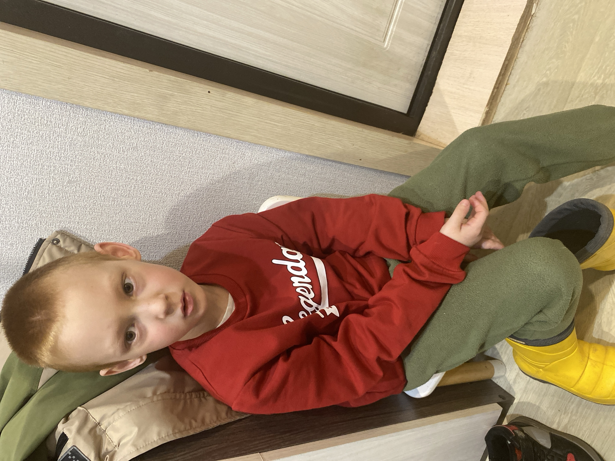 A young boy is sitting on a brown wooden shelf with a jacket underneath him. He is wearing a red sweatshirt with the word "legendary" on it and green sweatpants. His feet are clad in yellow rain boots. To the right is a pair of black shoes. The room is a hallway with light grey patterned wallpaper, a light wood door with a black frame, and light wood flooring. The location is Noviy, Russia.