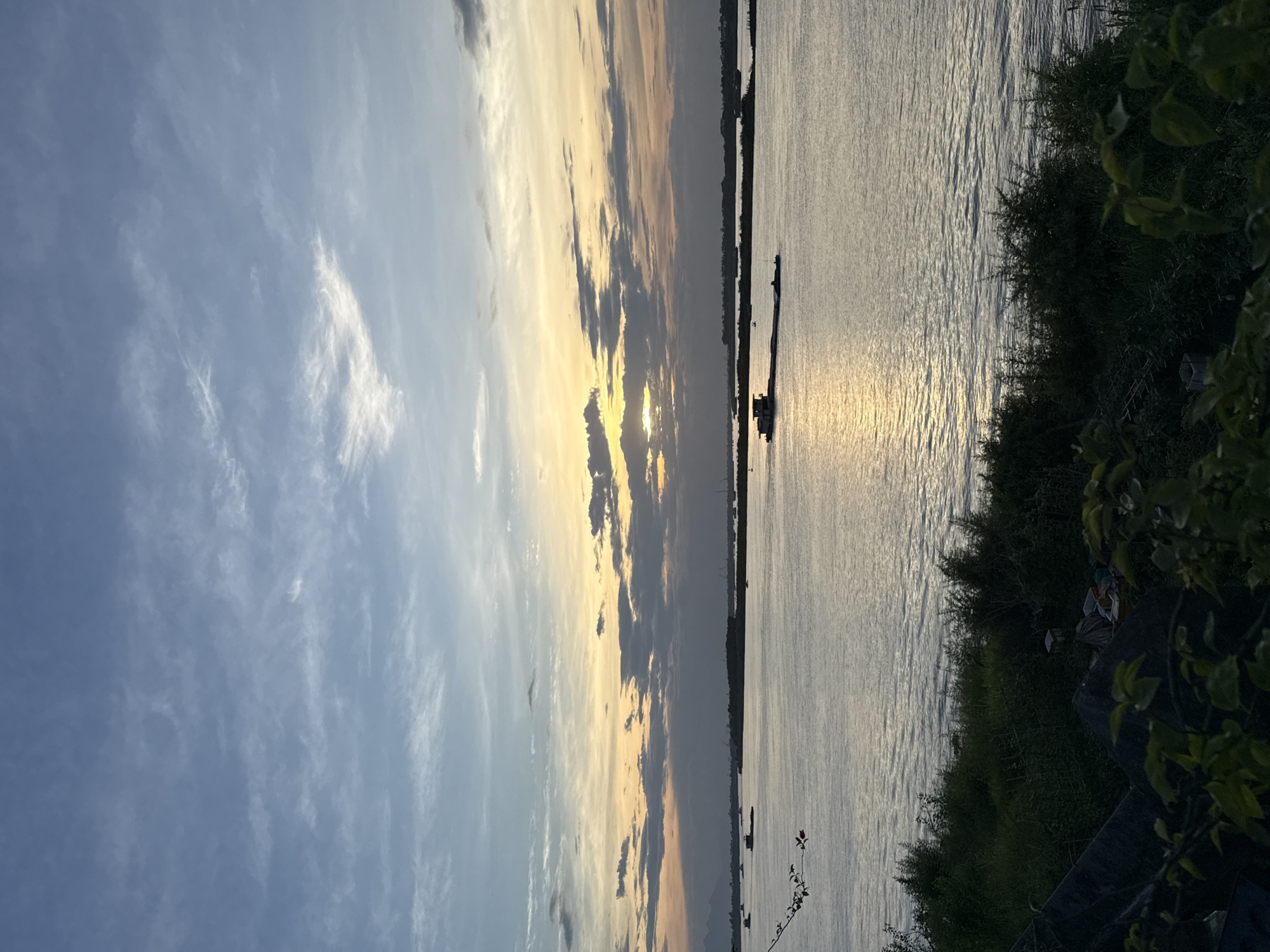 The image shows a body of water with foliage to the right and a sunset sky above. The sun is partially obscured by clouds. A long, dark shape is visible on the water. The location may be in or around Hà Nội, Vietnam, based on the image.