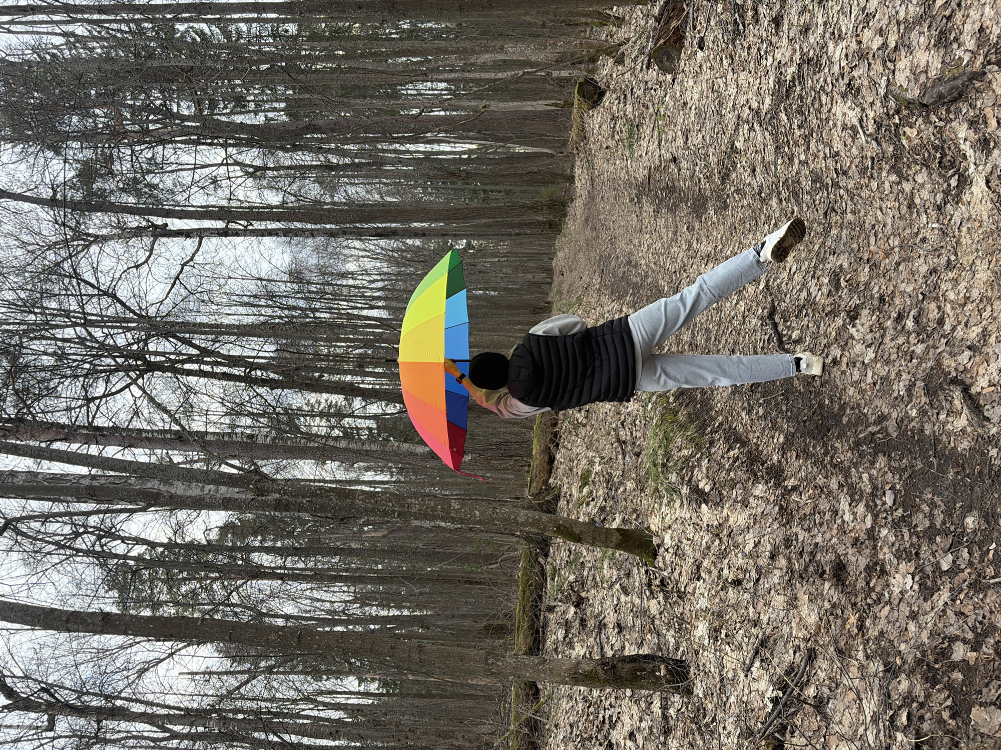 A person stands in a wooded area holding a colorful umbrella. The ground is covered with dead leaves. Bare trees surround the scene. The person wears gray sweatpants, white shoes, a black vest, and a black hat. The location is likely outside the city of Novyi, Russia given the wooded environment.