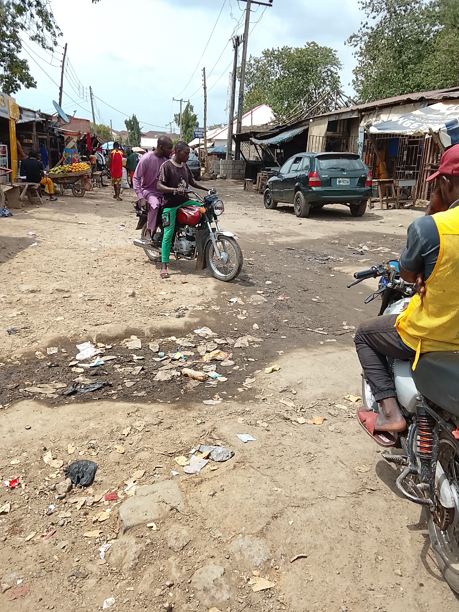 The image depicts a dirt road in Abuja, Nigeria, likely within a bustling market area. The road surface is uneven with patches of mud and scattered debris. Two men on a red motorcycle are in the middle ground, the driver wearing green pants and the passenger in a purple shirt. To the right, a man in a yellow vest is seated on another motorcycle. In the background, there is a green car parked next to informal storefronts with corrugated iron roofs. Other pedestrians and vendors are seen further up the road, along with a cart laden with produce. Power lines run overhead.