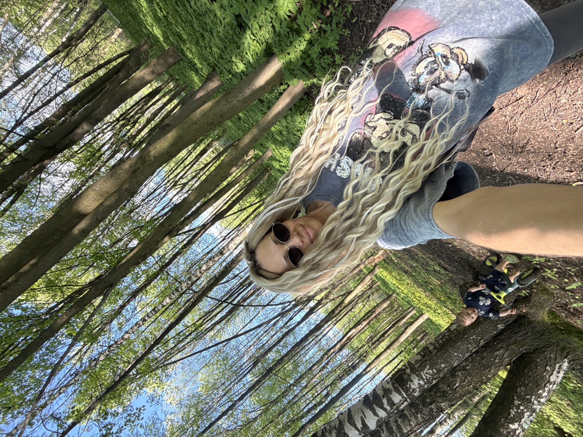 The image shows a woman and a child in a forest setting. The woman is wearing sunglasses and a printed shirt. The child is on a small bike. The background is filled with trees, and the sky is visible through the branches. Rusanovo, Russia, is not readily apparent from this image.
