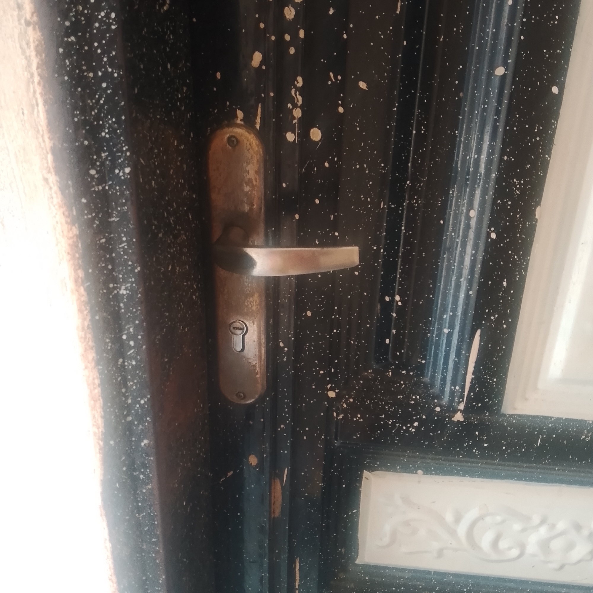 The image shows a close-up of a dark, speckled door with a metallic handle and lock. The door appears to be made of metal and has a weathered appearance with some scuff marks and paint chips. The handle is made of a shiny, worn metal, and below it is a keyhole. To the left of the door, a bright, out-of-focus light source is visible, possibly natural light from a window or doorway. The right side of the door shows a decorative white panel with embossed floral patterns. This scene likely depicts an exterior door in Jalingo, Nigeria, given the context provided.