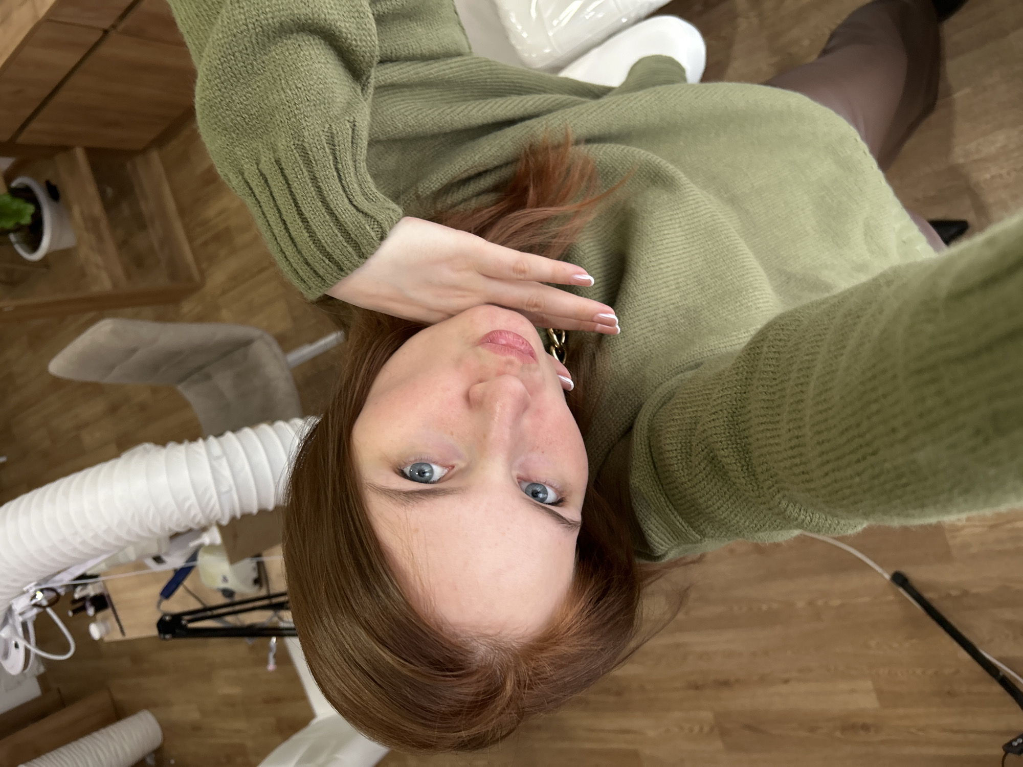 A woman with blue eyes and auburn hair is taking a selfie. She is wearing a green sweater and brown tights. The background includes wooden flooring, a white plastic tube, and equipment such as a camera stand. There is a white planter with a green plant. The location in the city of Chaikovsky, Russia, cannot be verified based on visual information in the image.