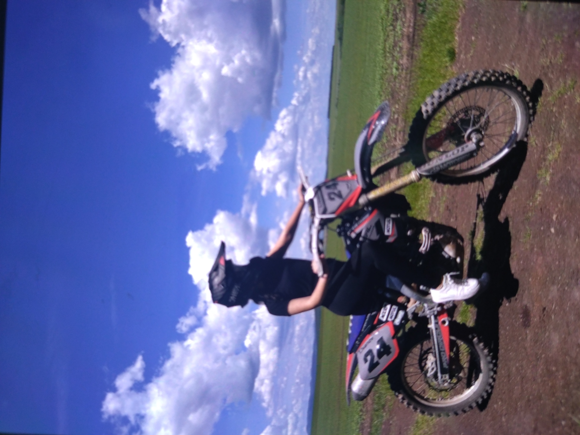 A person wearing a helmet, a dark shirt, dark pants and white sneakers is sitting on a motorcycle, appearing to be performing a wheelie. The motorcycle is blue and red and has the number 24 on the front. The background features a blue sky with white clouds, a green field, and what appears to be dirt ground. The location of the image is not visible in the photo.