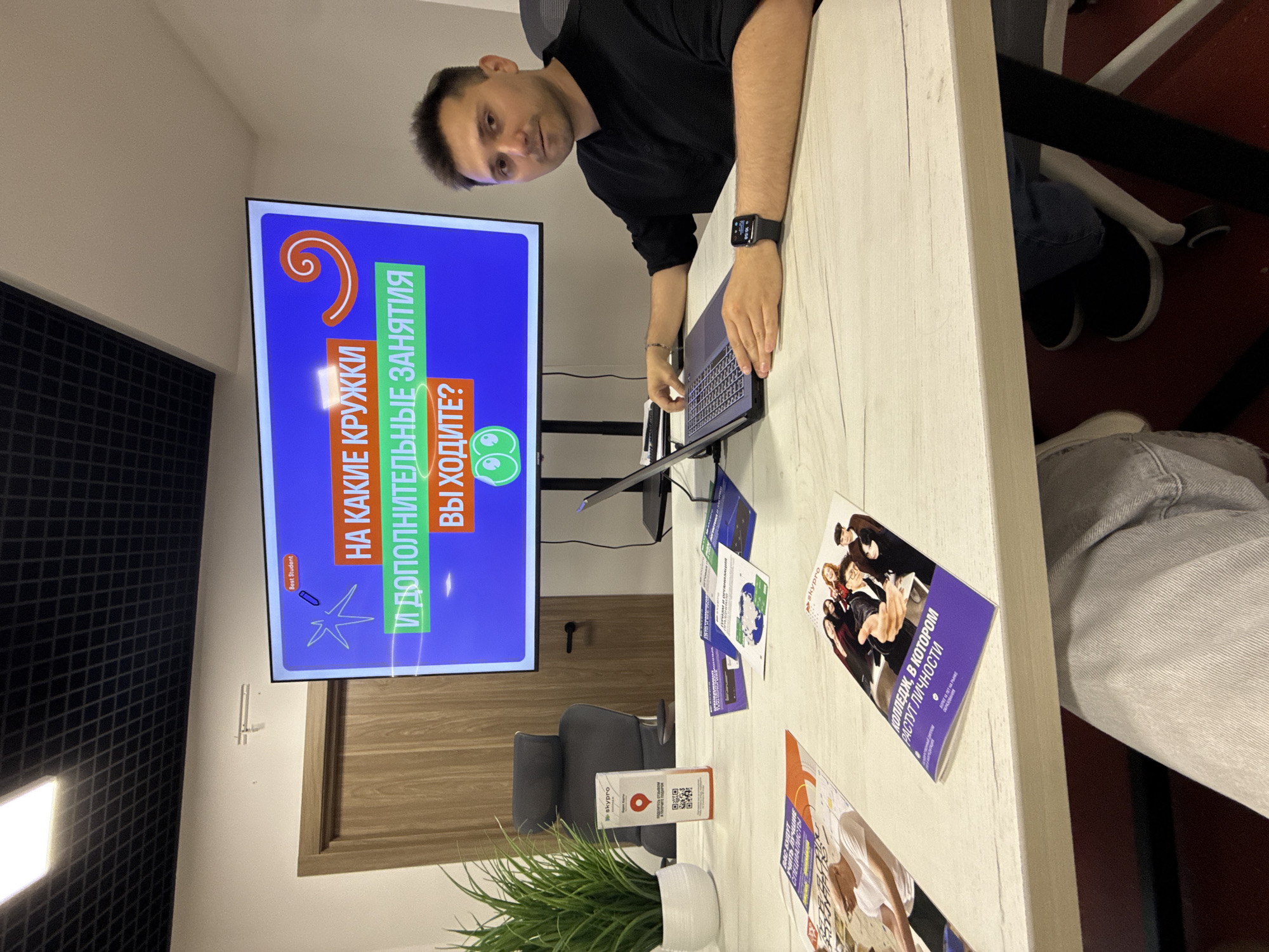 The image depicts a man sitting at a table with a laptop, in what appears to be an office or classroom setting. A large screen behind him displays text in Russian, which translates to "What kind of clubs and extracurricular activities are you interested in?". There are also several brochures and a plant on the table. The man is wearing a black shirt and a watch on his left wrist. The background shows a door and part of a wall with a ceiling light. There is no discernible information in the image to suggest the location is the city of Sochi, Russia.