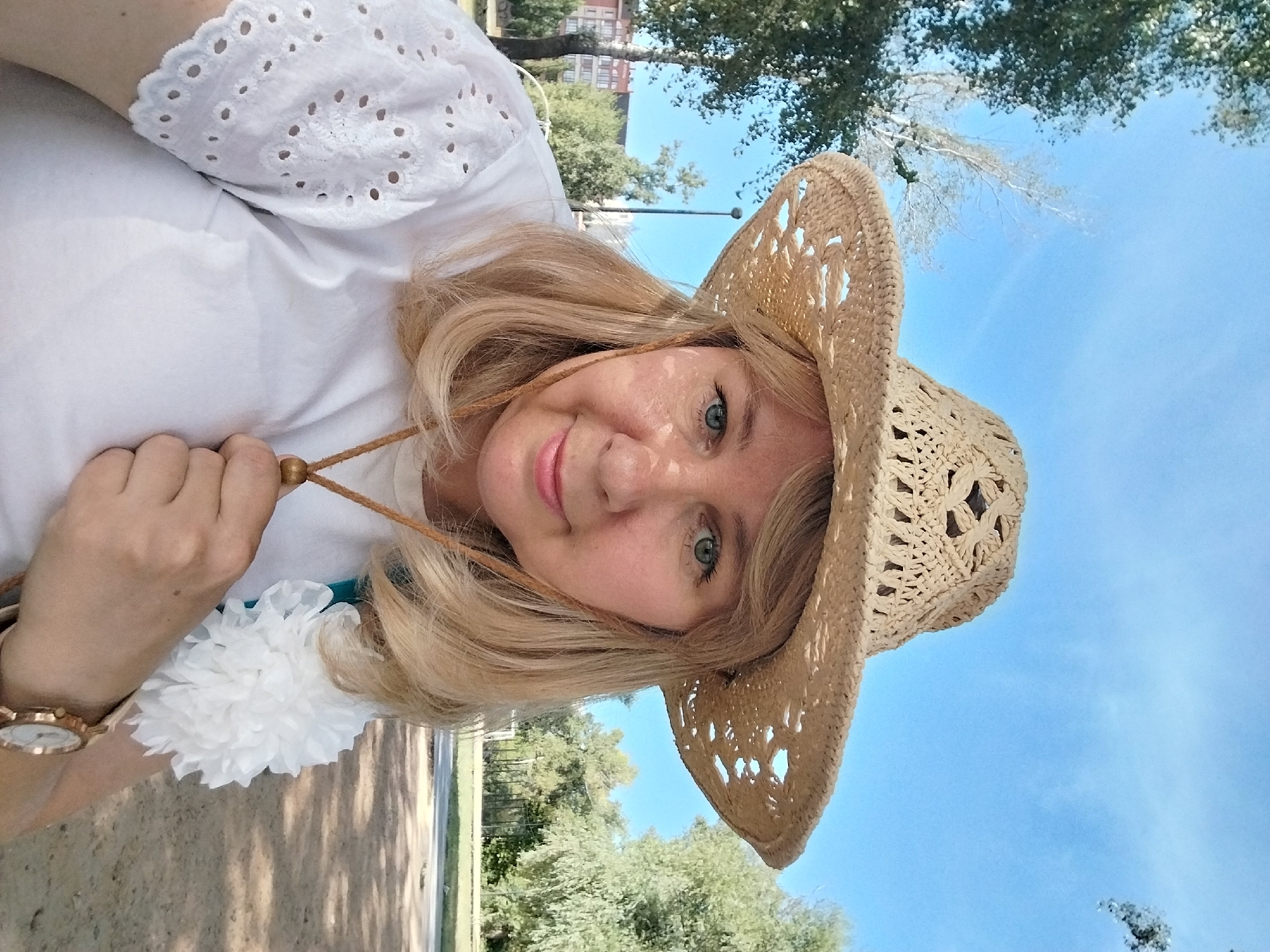 A woman wearing a straw cowboy hat and a white eyelet shirt poses for a selfie outdoors. She has blonde hair, blue eyes, and is smiling. A white floral accessory is attached to her shirt. She is wearing a wristwatch. The background shows trees, buildings, and a blue sky. The image was taken in Orenburg, Russia.