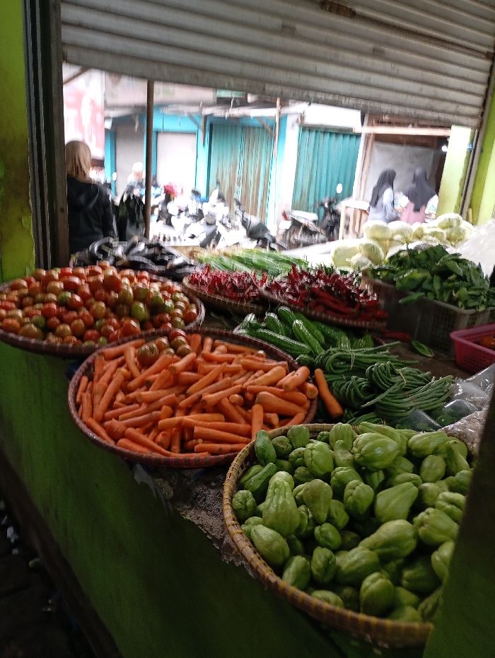 The image shows a street market in Kutawaringin, Indonesia, located at coordinates -6.953 latitude and 107.524 longitude. Various vegetables are displayed in baskets, including tomatoes, carrots, cucumbers, green beans, and chayote squash. The scene is outside, likely under a covered area, with several motorbikes parked in the background. There are several people standing or walking in the background. The image captures a lively scene of a bustling marketplace.