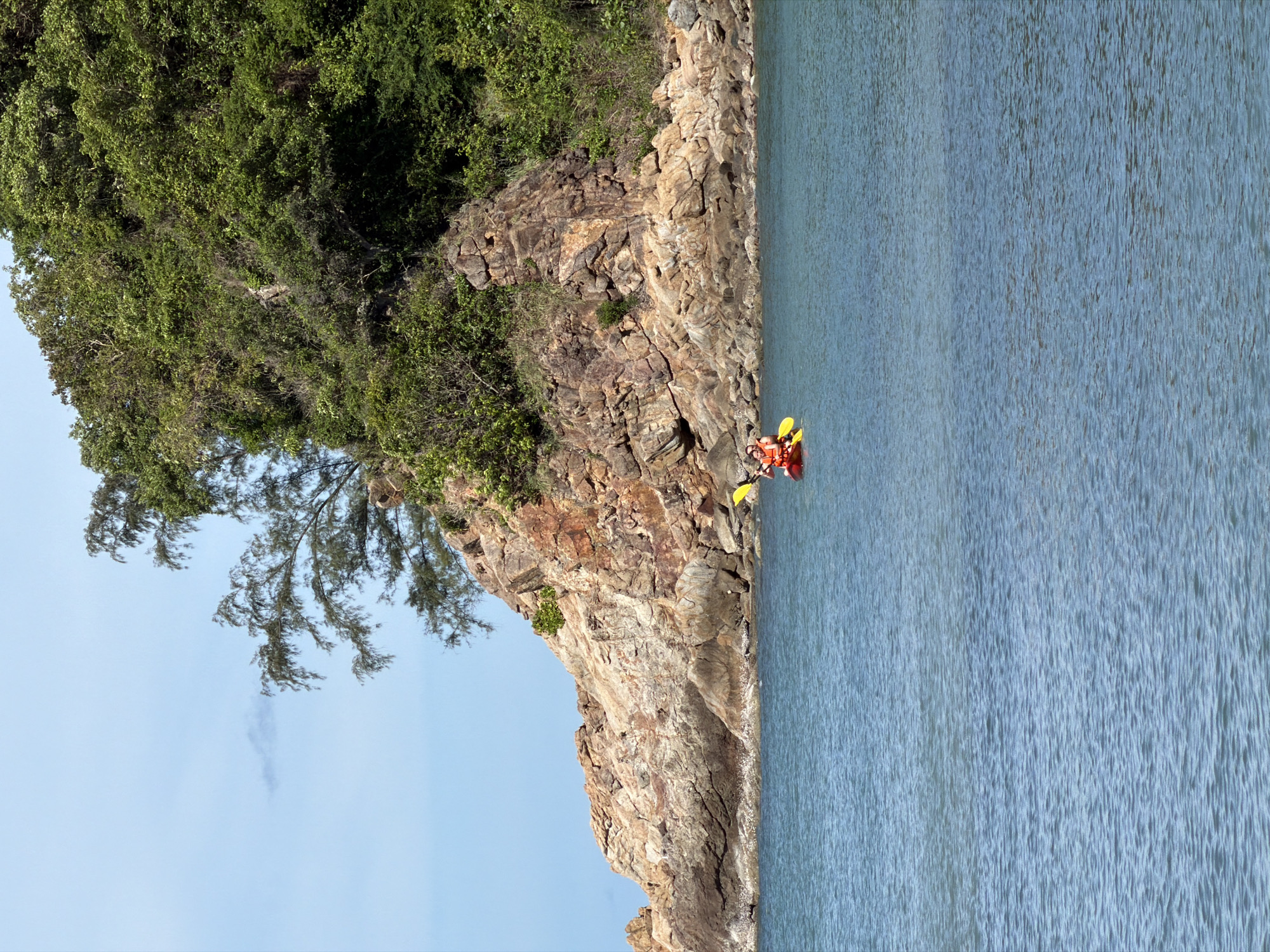 This is a vertical, medium-shot photograph taken outdoors. The scene features a rocky cliff face covered in lush green vegetation on the left, bordering a calm, light blue body of water on the right. The sky, a pale blue, occupies the upper left portion of the image, with some wispy white clouds visible.
In the water, near the base of the cliff, a red kayak is visible with two people inside. They are wearing orange life vests. One person is paddling with a yellow paddle, and another is seated behind them. The kayak is positioned close to the rocky shore.
The lighting suggests it is daytime, likely late morning or early afternoon, with clear weather. The overall mood is peaceful and serene, highlighting the natural beauty of the coastal landscape. The location is identified as Baan Plai Laem, Thailand. There is no visible text in the image.