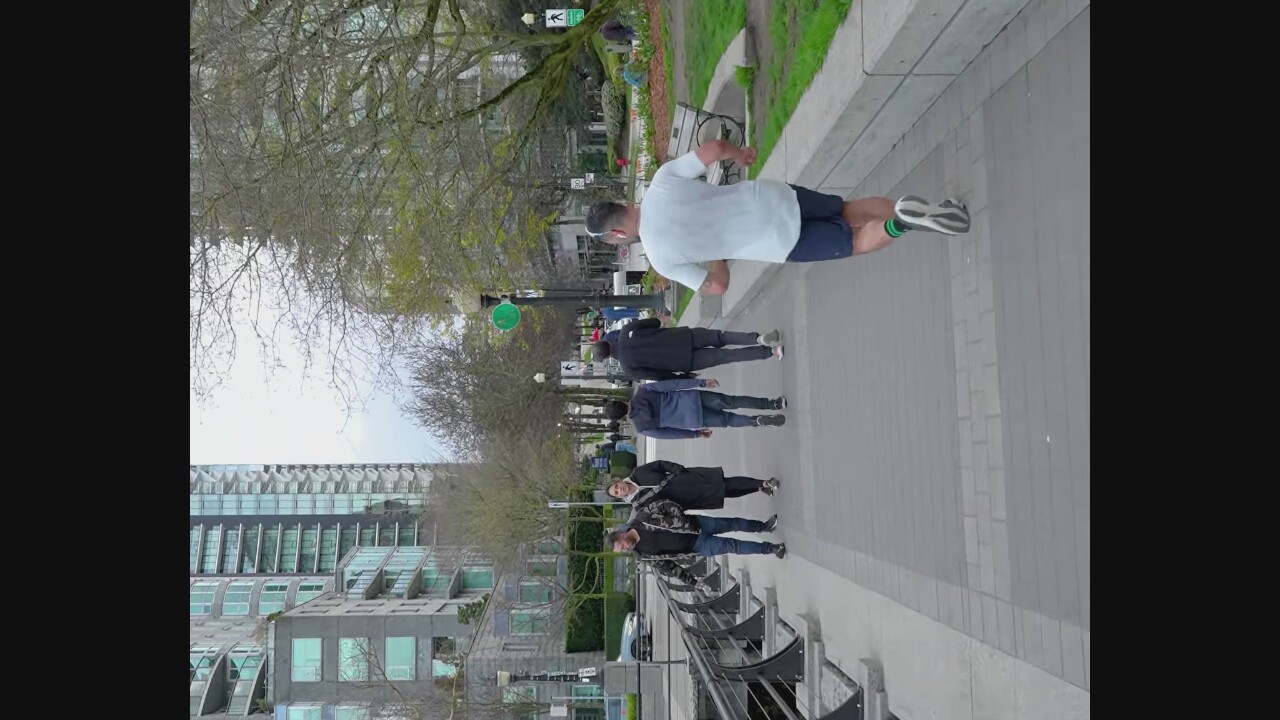 A group of five people are walking and running along a paved pathway next to a body of water in Vancouver, Canada. In the foreground, a man is running away from the camera, wearing a white t-shirt, dark shorts, and striped socks. Behind him, four people are walking away from the camera on the same pathway. From left to right, they are a man in a camouflage jacket and jeans, a woman in a black coat, a young man in a blue hoodie and jeans, and a man in a black jacket and pants. The pathway is lined with a decorative railing on the left and a concrete barrier and grassy area with trees on the right. In the background, modern high-rise buildings with glass facades dominate the skyline. The sky is overcast.