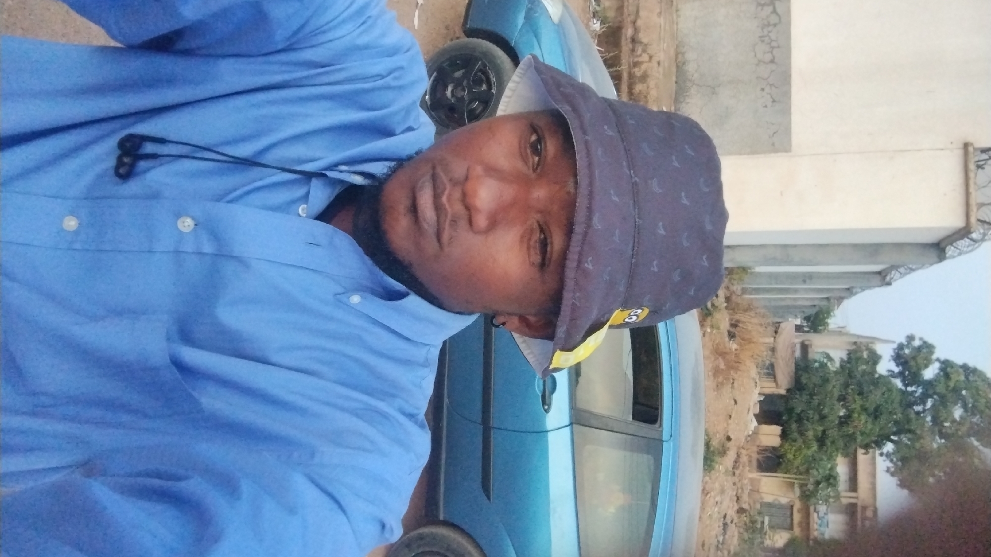 A man wearing a blue button-down shirt and a patterned bucket hat stands in front of a light blue car. He has earphones around his neck. The background features a building with a concrete structure and a tree. The location appears to be an urban area, likely in Jos, Nigeria.