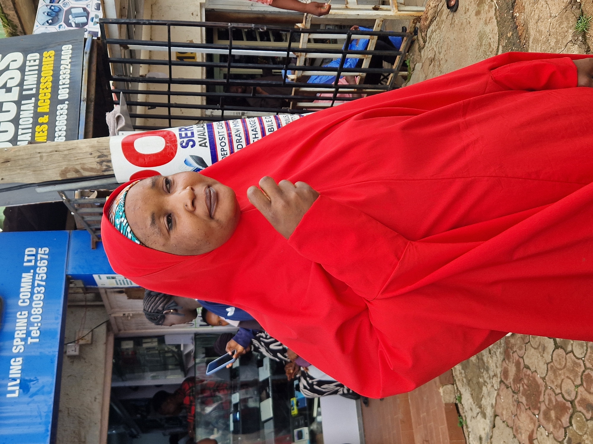 This is a medium shot of a woman wearing a red headscarf and dress, standing in front of a storefront. She is looking towards the camera with a neutral expression. Behind her, a metal gate separates the storefront from the street. Several signs are visible, including one that reads "SUCCESS NATIONAL LIMITED PHONES & ACCESSORIES" and another that says "LIVING SPRING COMM. LTD Tel: 08093756675". Other signs are partially obscured but appear to be advertisements. Inside the storefront, electronic devices are displayed. The overall scene appears to be an outdoor market or a street vendor setting. The lighting suggests it is daytime, but no specific weather conditions are discernible. The woman's clothing and the storefront suggest a location in a region where headscarves are common.