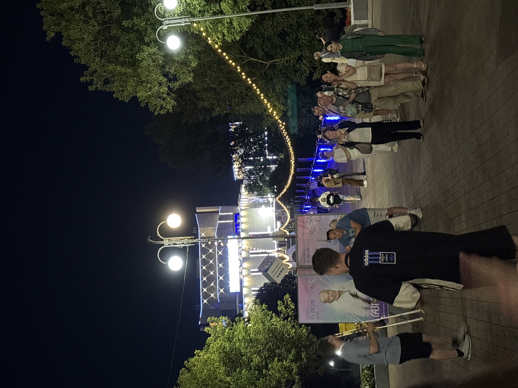 This is a nighttime outdoor scene in Vitebsk, Belarus, likely on a pedestrian walkway or plaza. In the foreground, a man in a black Adidas t-shirt with white sleeves faces away from the camera, obscuring the view of other people. To his left, a man plays a brass instrument, possibly a trombone, while wearing a grey t-shirt and black shorts.
Behind them, a large poster is displayed, featuring portraits of two men and text in Russian, possibly an advertisement for an event on July 15th. Further in the background, a modern building is illuminated with numerous lights, creating a festive atmosphere. String lights with yellow bulbs are strung across the scene, adding to the ambiance. Several other individuals are visible in the midground and background, some walking and some standing and observing. The ground is paved with brick-like stones. Trees with green foliage are present on the sides of the scene. The sky is dark, indicating it is nighttime.